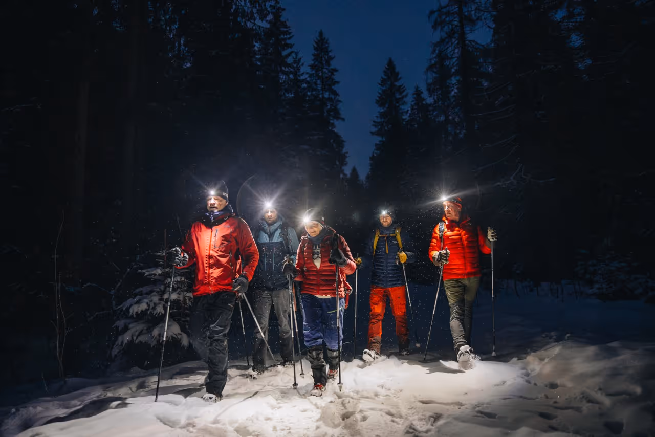 A group of people walking through a snow-covered forest at twilight, with headlamps