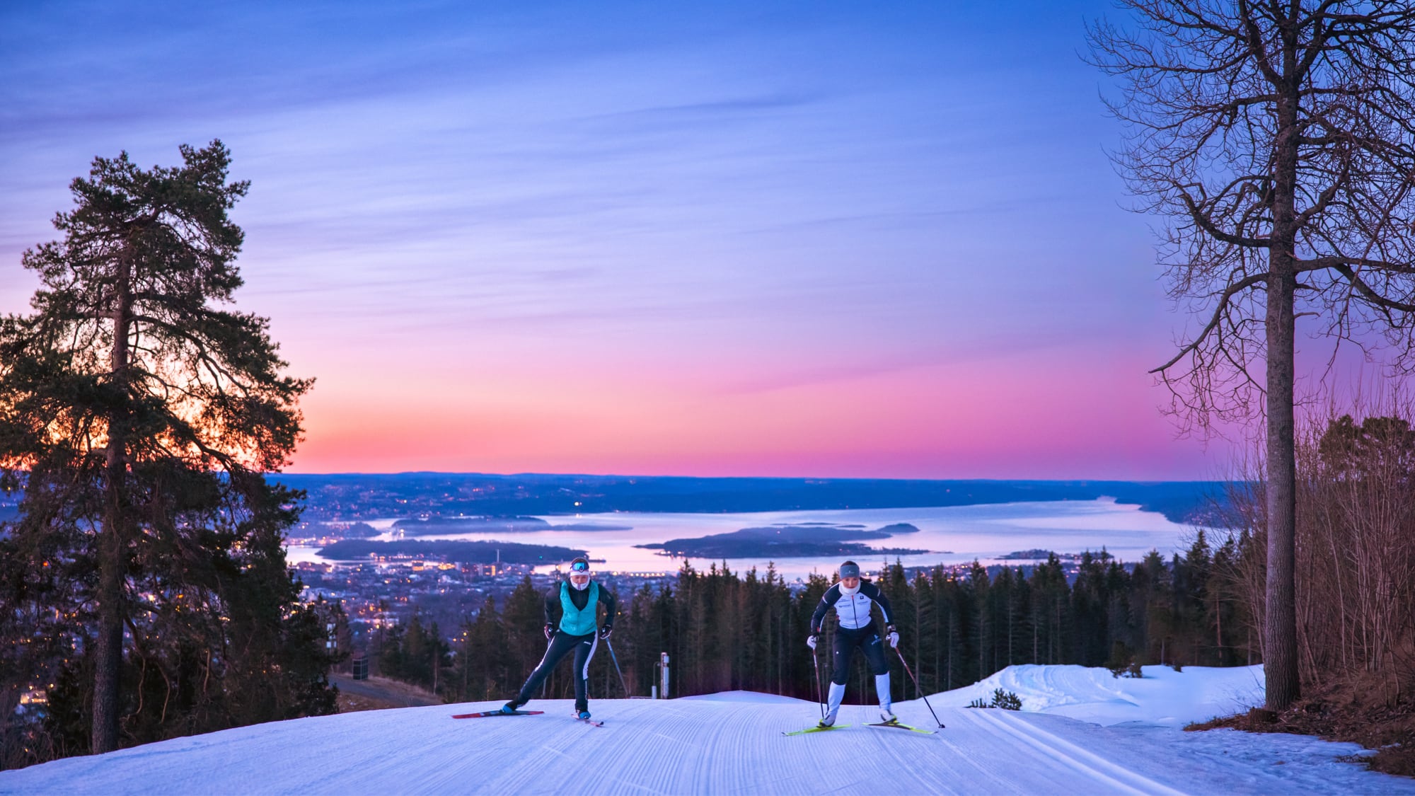 Two cross-country skiers in red outfits skiing at Holmenkollen/Tryvann with a scenic view of Oslo and the fjord in the background during sunset.
