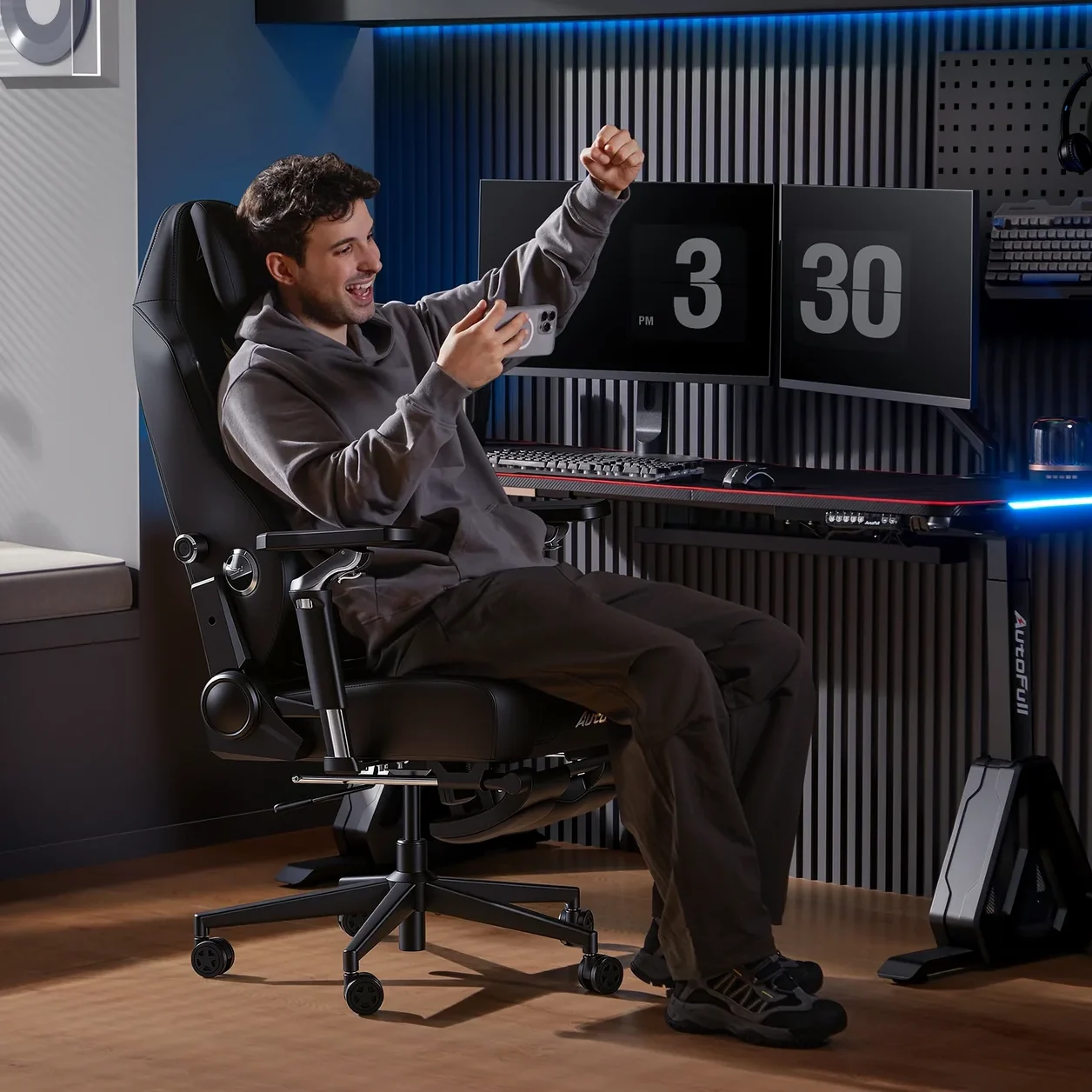 Man in gray hoodie sitting in a black ergonomic gaming chair from AutoFull, smiling and raising his fist in front of a dual-monitor desk.