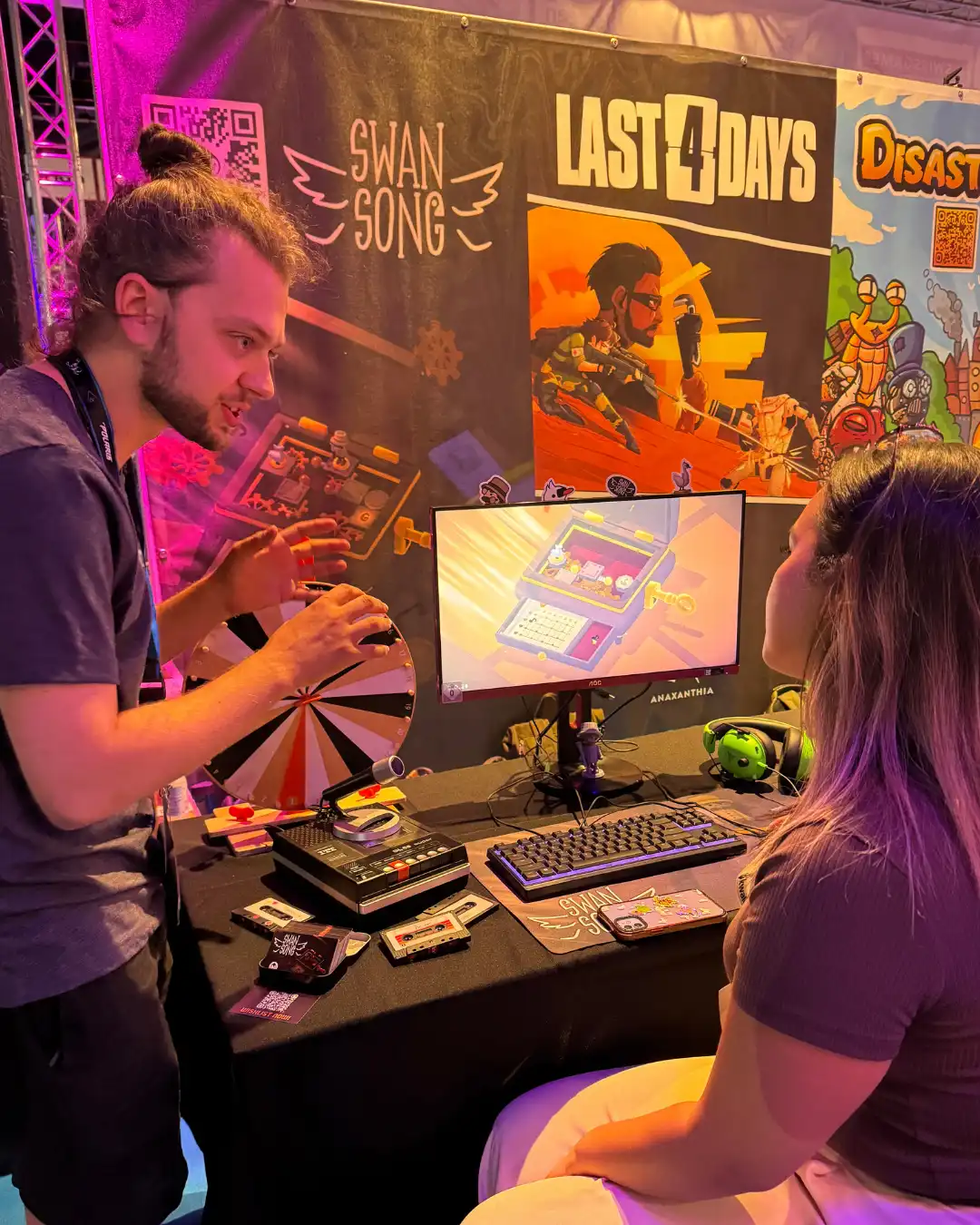 Photograph of the Swan Song booth at amescom. A lady sits at the PC playing the demo, while the developer for the game animatedly explains. On the desk is a keyboard & monitor with Swan Song, & a tape recorder to record messages.