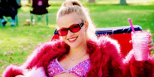 GIF image from the movie, Legally Blonde. Elle, a blonde woman, is smiling at the camera while seated in a pink lounge chair. She is wearing a pink sparkle bikini, a fluffy pink jacket, & pink sunglasses. She is holding up a pink cup & straw in her right hand.