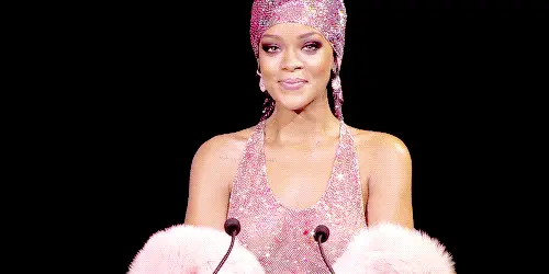 GIF image of Rihanna wearing a pink crystal gown & durag. She is smiling & giggling into a microphone.