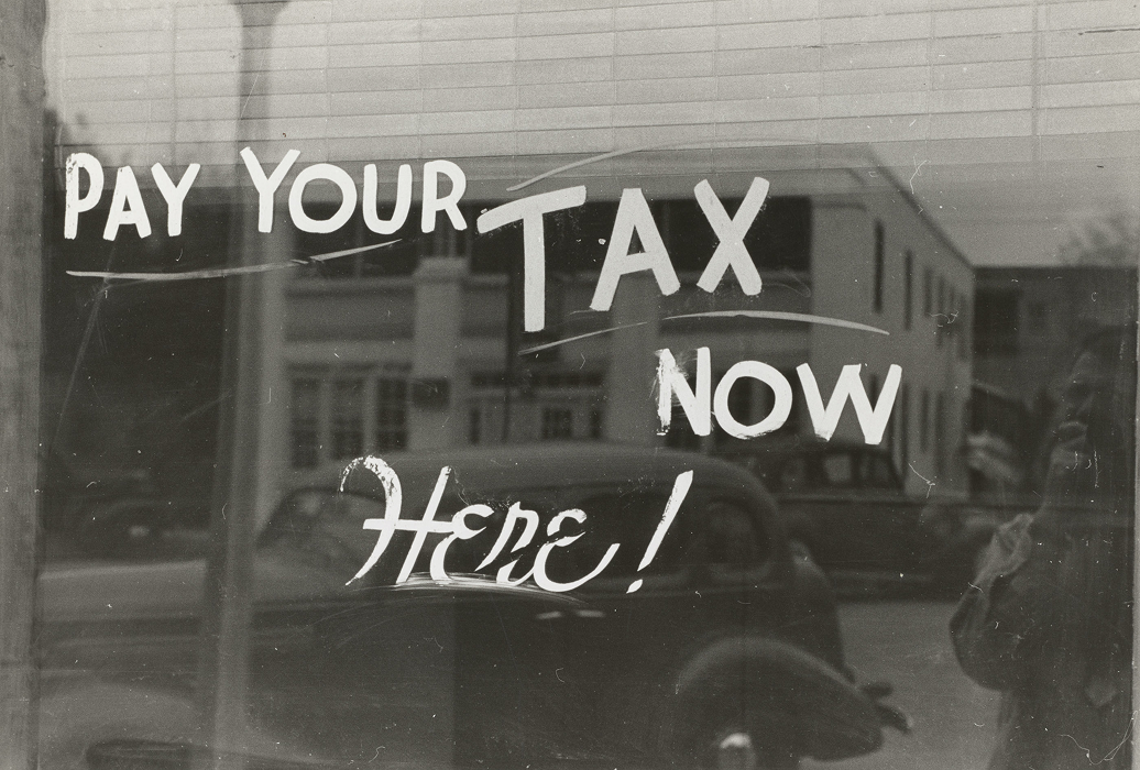 Vintage black and white storefront window sign reading "Pay Your Tax Now Here!" reflecting a street scene.