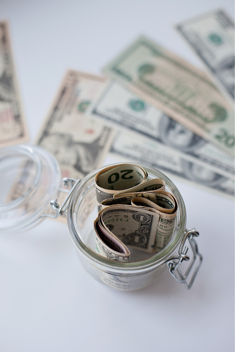 Glass jar filled with rolled up dollar bills with additional cash spread out on a white surface behind it