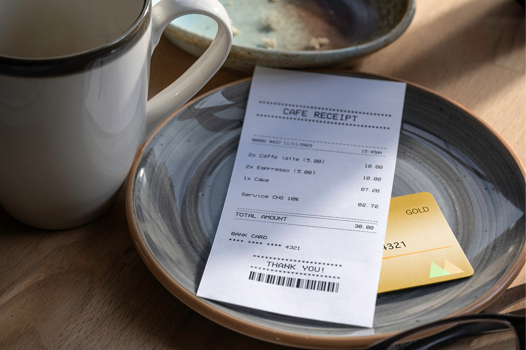 Cafe receipt showing itemized charges on a ceramic plate next to a gold credit card and a white coffee mug on a wooden table