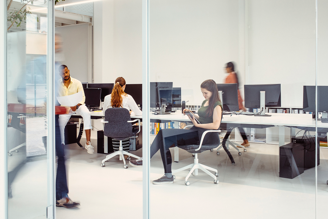 Busy modern office viewed through glass walls with employees working at desks, one standing reviewing documents with a colleague