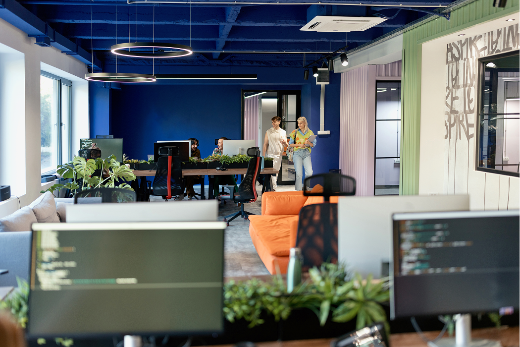 Modern open-plan office with blue accent walls, indoor plants, orange sofa, computer workstations, and two employees walking and talking