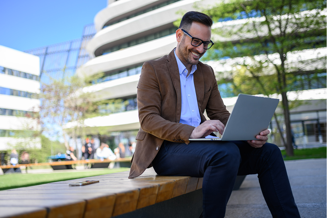 Smiling businessman in a brown blazer and glasses working on a laptop while seated on an outdoor bench in front of a modern office building