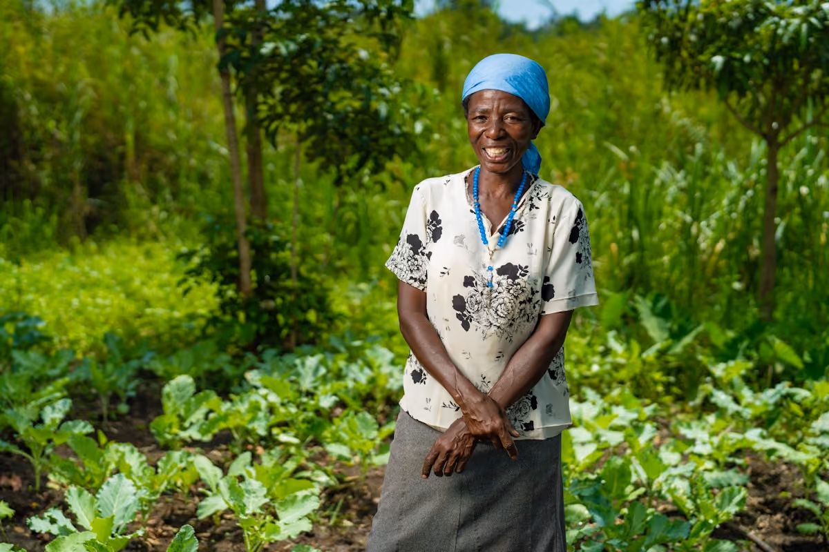 A woman wearing a teal headscarf smiles while standing in front of thriving farm in Kenya.