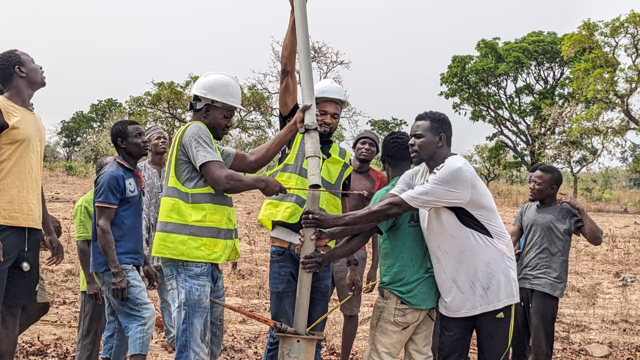 A group of men working together outdoors to install or secure a vertical metal pole in the ground, with two men wearing reflective safety vests and helmets while others assist around them.