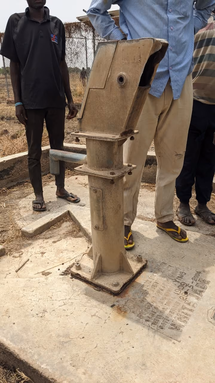Metal water handpump installed on a cement platform with three men standing nearby, one wearing a blue shirt and beige pants with yellow sandals, outdoors on dry ground with a wire fence in the background.