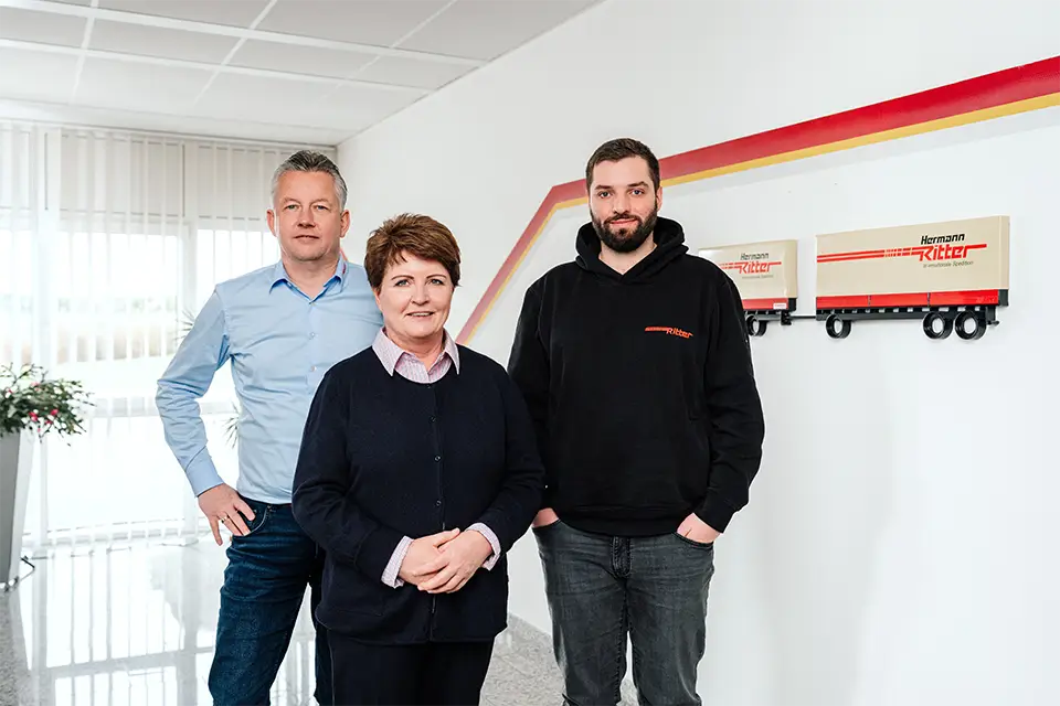 Drei Personen stehen nebeneinander in einem hellen Büro mit weißen Wänden und drei Modell-LKWs an der Wand, die den Schriftzug 'Herrmann Ritter' tragen.