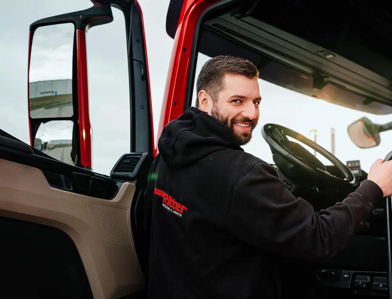 Hermann Ritter trägt einen schwarzen Kapuzenpullover mit rotem Schriftzug 'Ritter', besteigt die Fahrerkabine eines roten Lkw und schaut lächelnd zur Kamera.