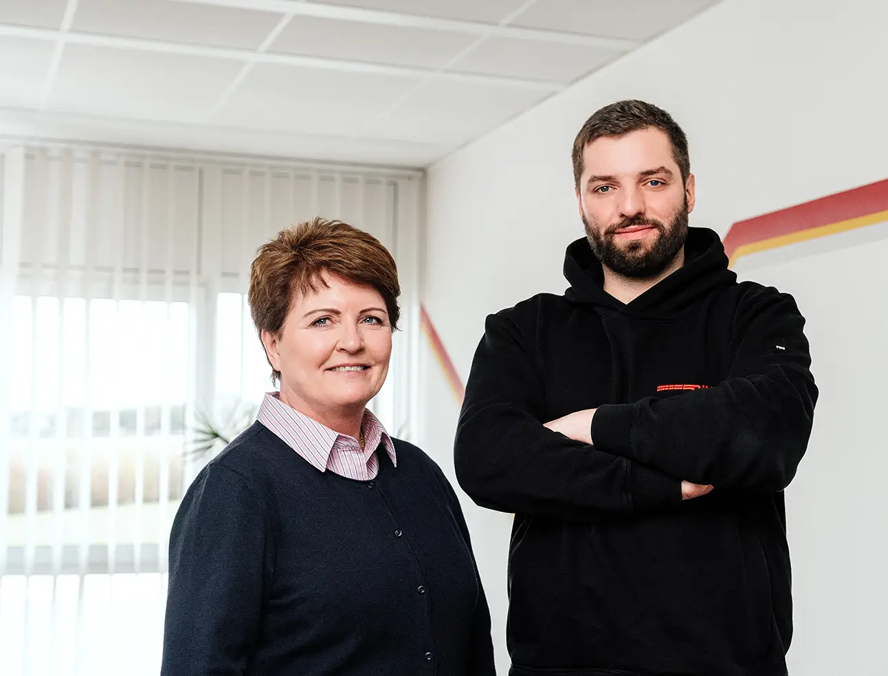 Pamela und Hermann Ritter stehen in einem hellen Raum, er trägt einen schwarzen Kapuzenpullover, sie einen dunklen Pullover über einem Hemd.