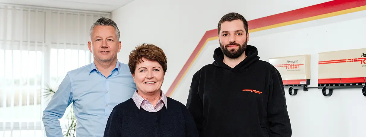 Drei Personen stehen nebeneinander in einem hellen Büro mit weißen Wänden und drei Modell-LKWs an der Wand, die den Schriftzug 'Herrmann Ritter' tragen.
