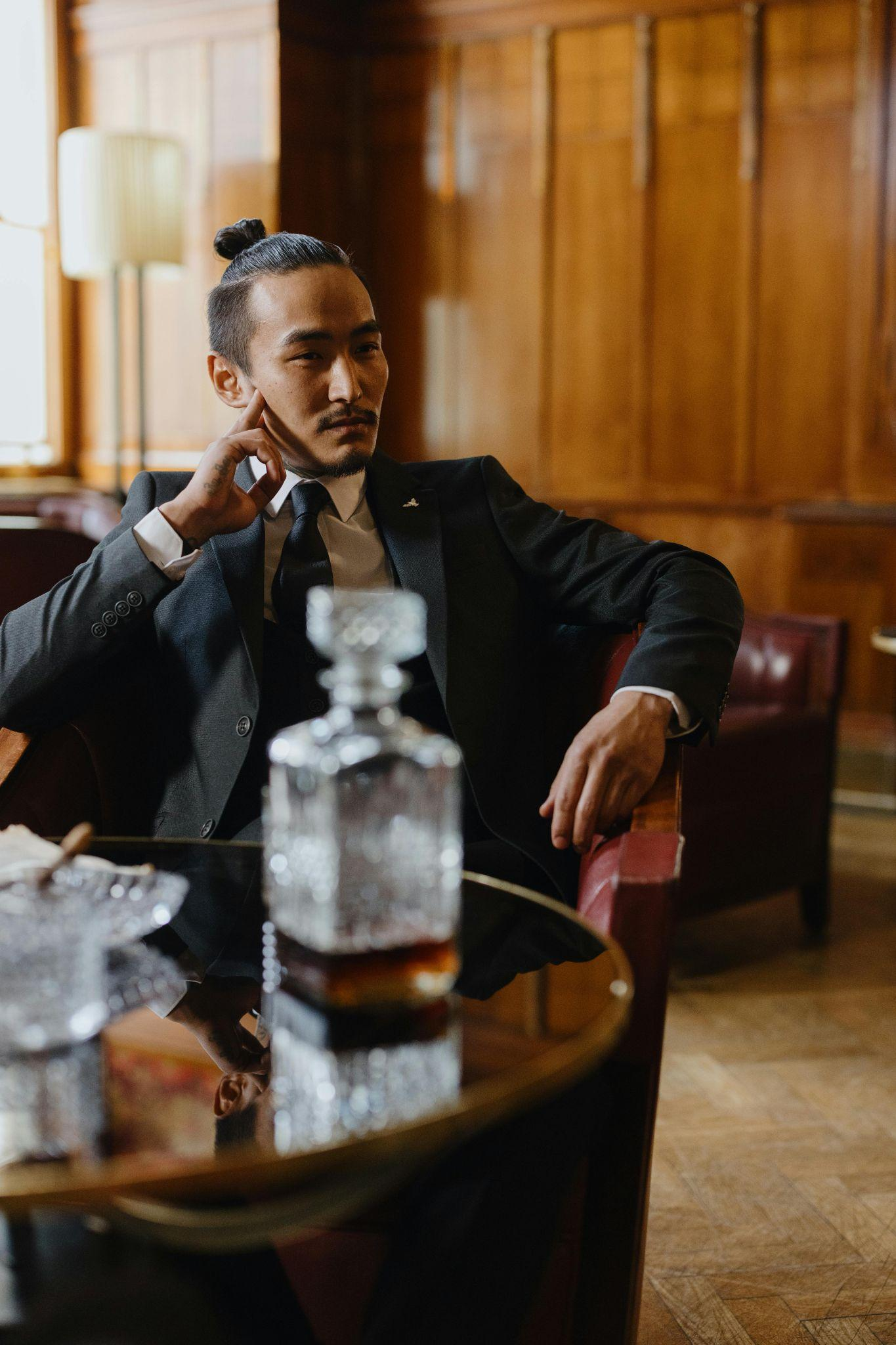 A man in a suit sitting in a leather chair, with a whiskey decanter on the table.