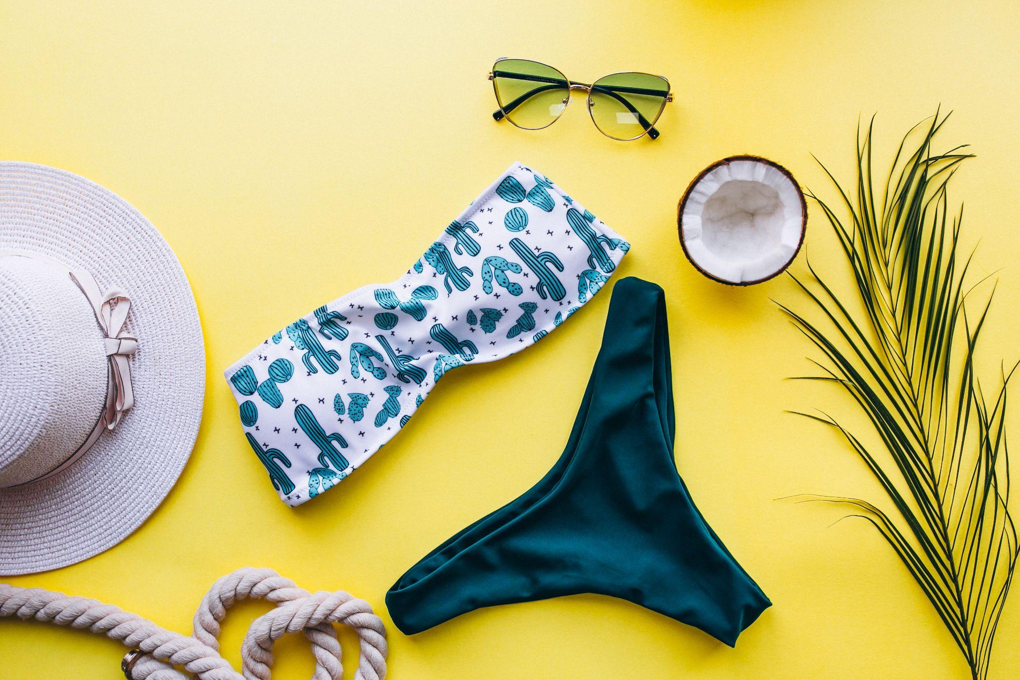 A flat lay of a bikini set, sunhat, sunglasses, and beach accessories on a yellow background.