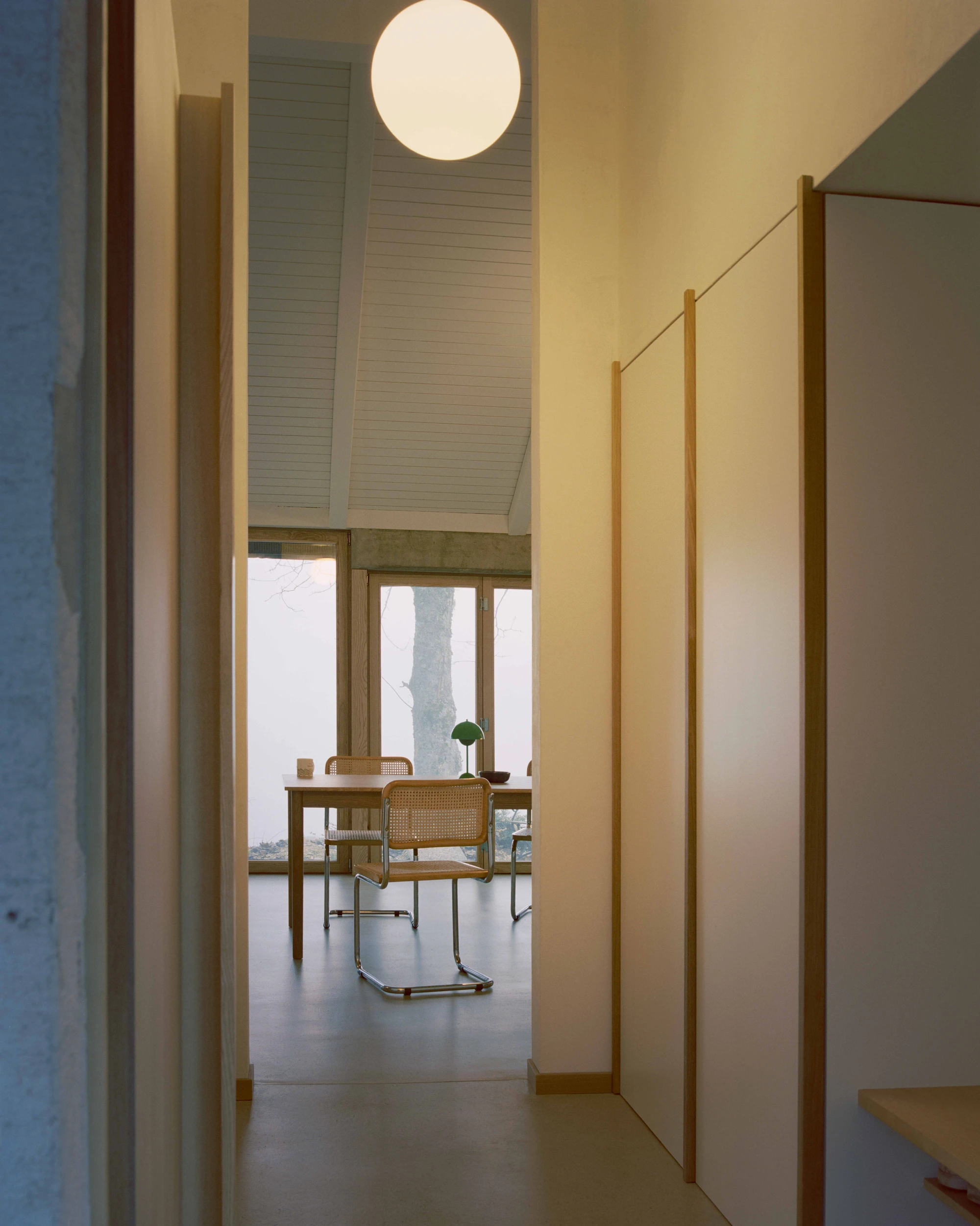 Modest white single-story villa with dark tile roof framed by bare winter trees in morning fog, autumn leaves scattered across a green lawn in the Swiss Jura foothills, Family Villa by Noue Studio, interior design