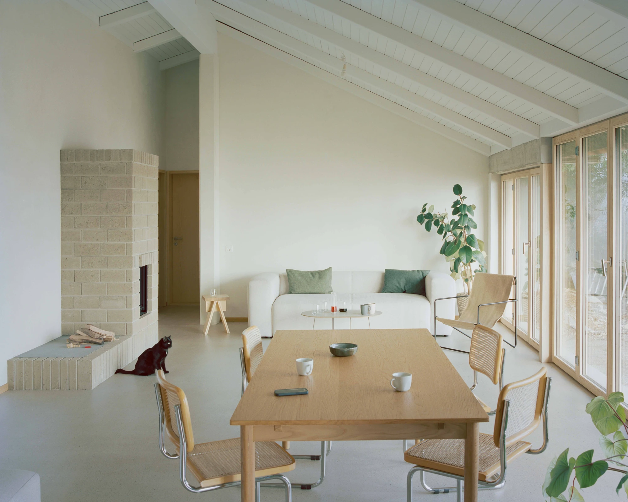 Modest white single-story villa with dark tile roof framed by bare winter trees in morning fog, autumn leaves scattered across a green lawn in the Swiss Jura foothills, Family Villa by Noue Studio, interior design