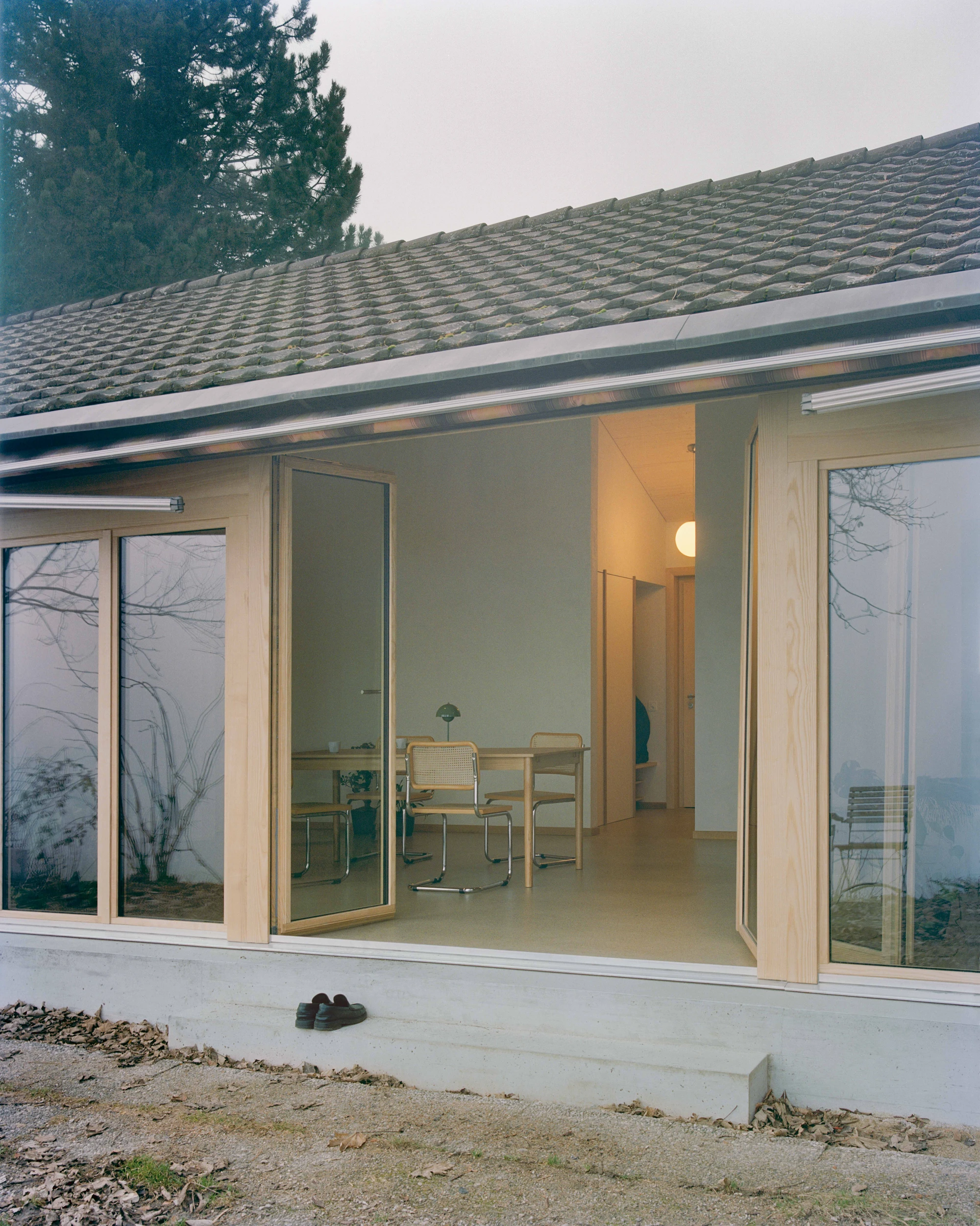 Modest white single-story villa with dark tile roof framed by bare winter trees in morning fog, autumn leaves scattered across a green lawn in the Swiss Jura foothills, Family Villa by Noue Studio, interior design