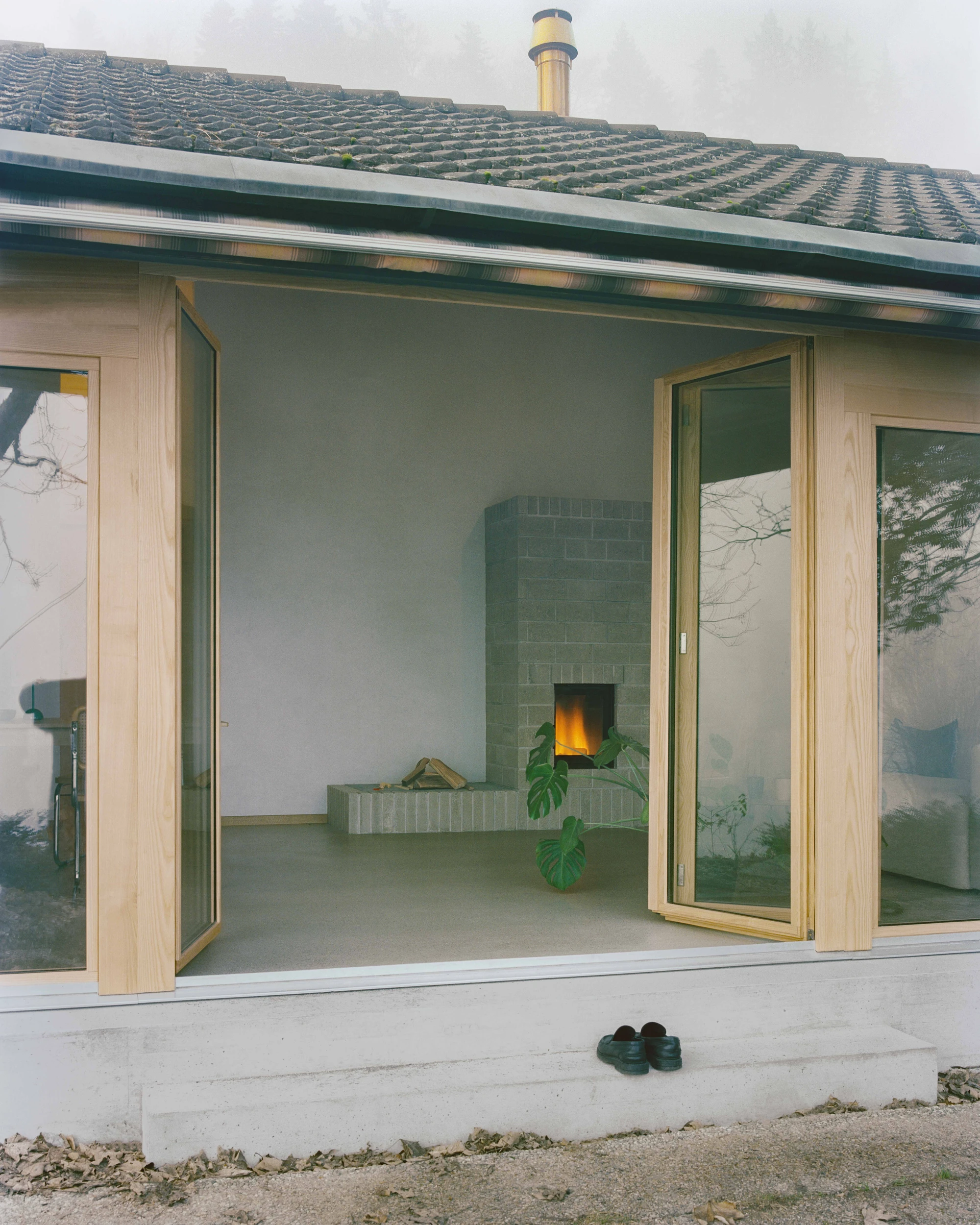 Modest white single-story villa with dark tile roof framed by bare winter trees in morning fog, autumn leaves scattered across a green lawn in the Swiss Jura foothills, Family Villa by Noue Studio, interior design