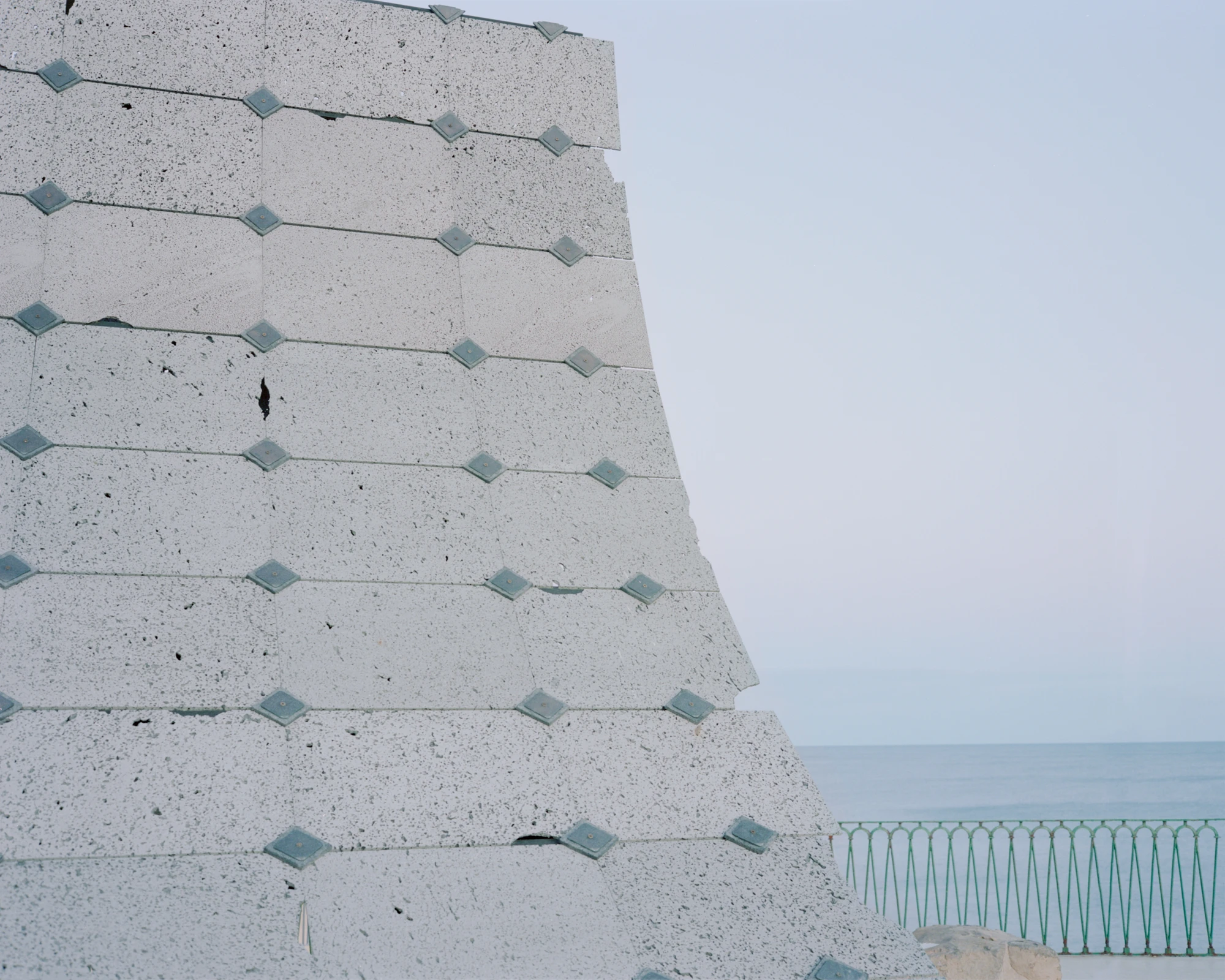 Pantalica Speculations by Leopold Banchini, An asymptotic timber and stone pavilion on a seaside promenade overlooking the Mediterranean, dark wood frame supporting stone-tile roof on massive limestone blocks