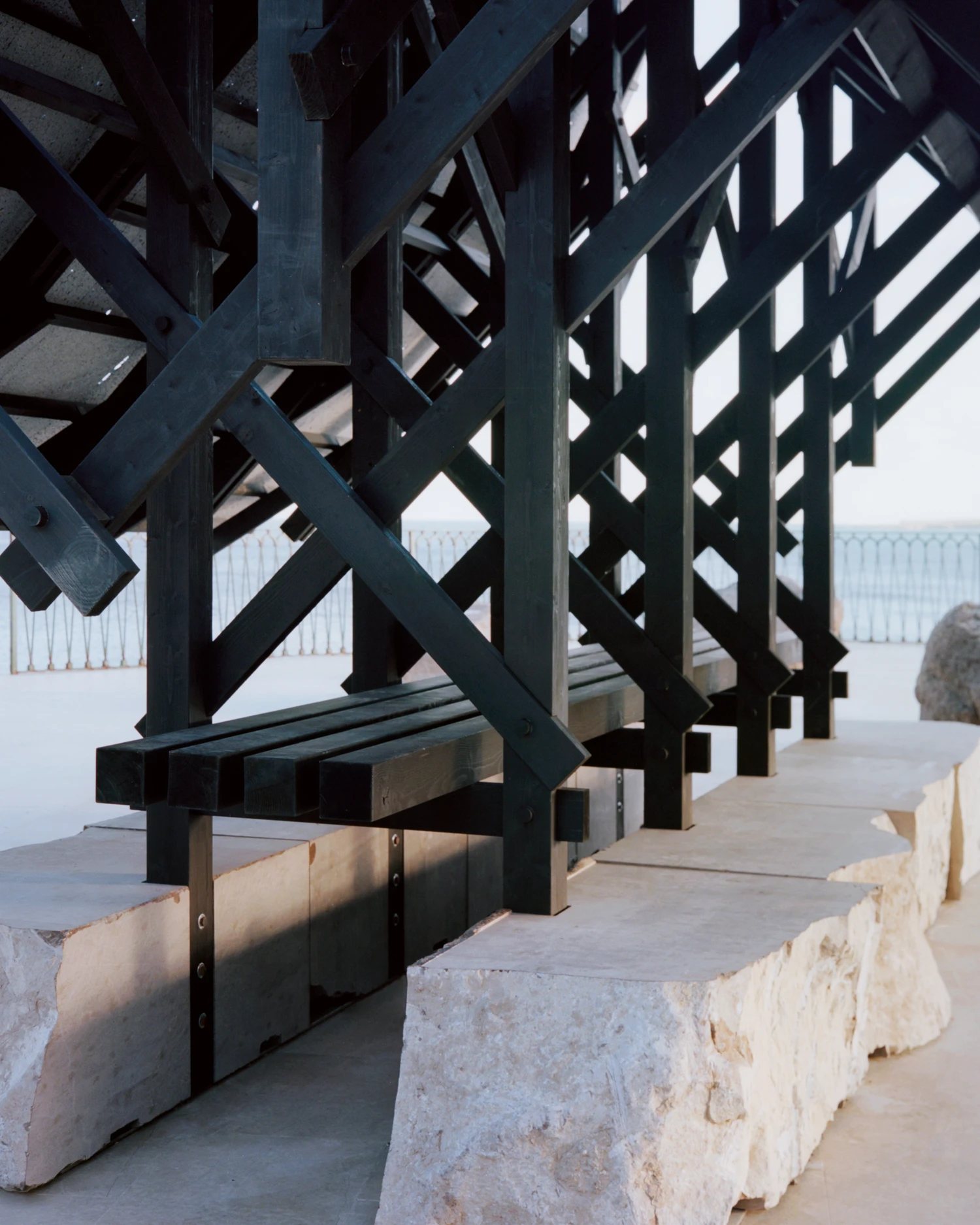 Pantalica Speculations by Leopold Banchini, An asymptotic timber and stone pavilion on a seaside promenade overlooking the Mediterranean, dark wood frame supporting stone-tile roof on massive limestone blocks