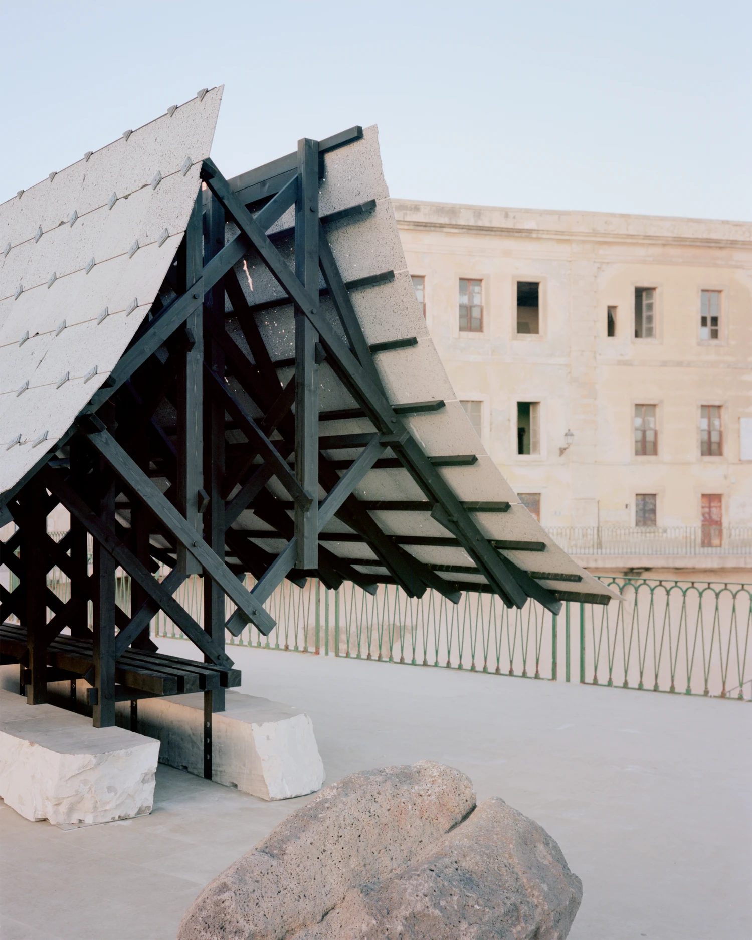 Pantalica Speculations by Leopold Banchini, An asymptotic timber and stone pavilion on a seaside promenade overlooking the Mediterranean, dark wood frame supporting stone-tile roof on massive limestone blocks