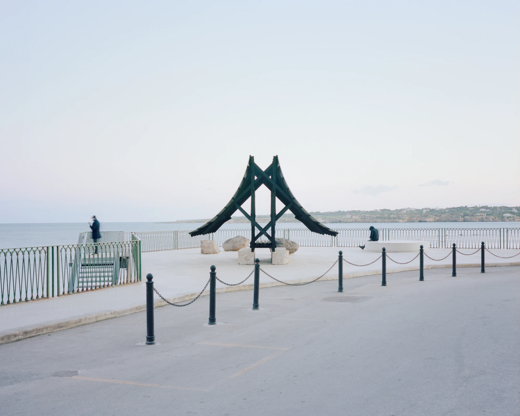Pantalica Speculations by Leopold Banchini, An asymptotic timber and stone pavilion on a seaside promenade overlooking the Mediterranean, dark wood frame supporting stone-tile roof on massive limestone blocks