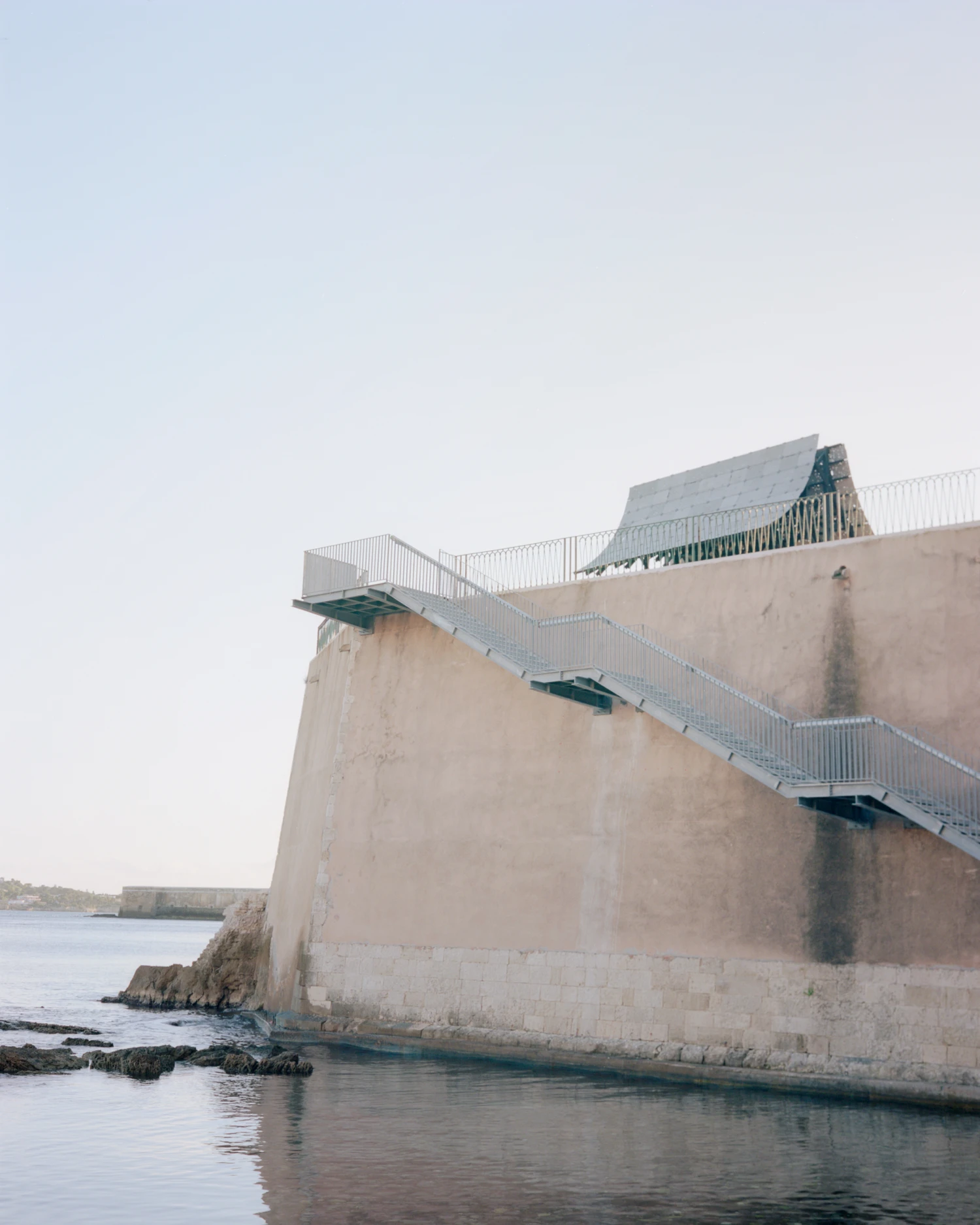 Pantalica Speculations by Leopold Banchini, An asymptotic timber and stone pavilion on a seaside promenade overlooking the Mediterranean, dark wood frame supporting stone-tile roof on massive limestone blocks
