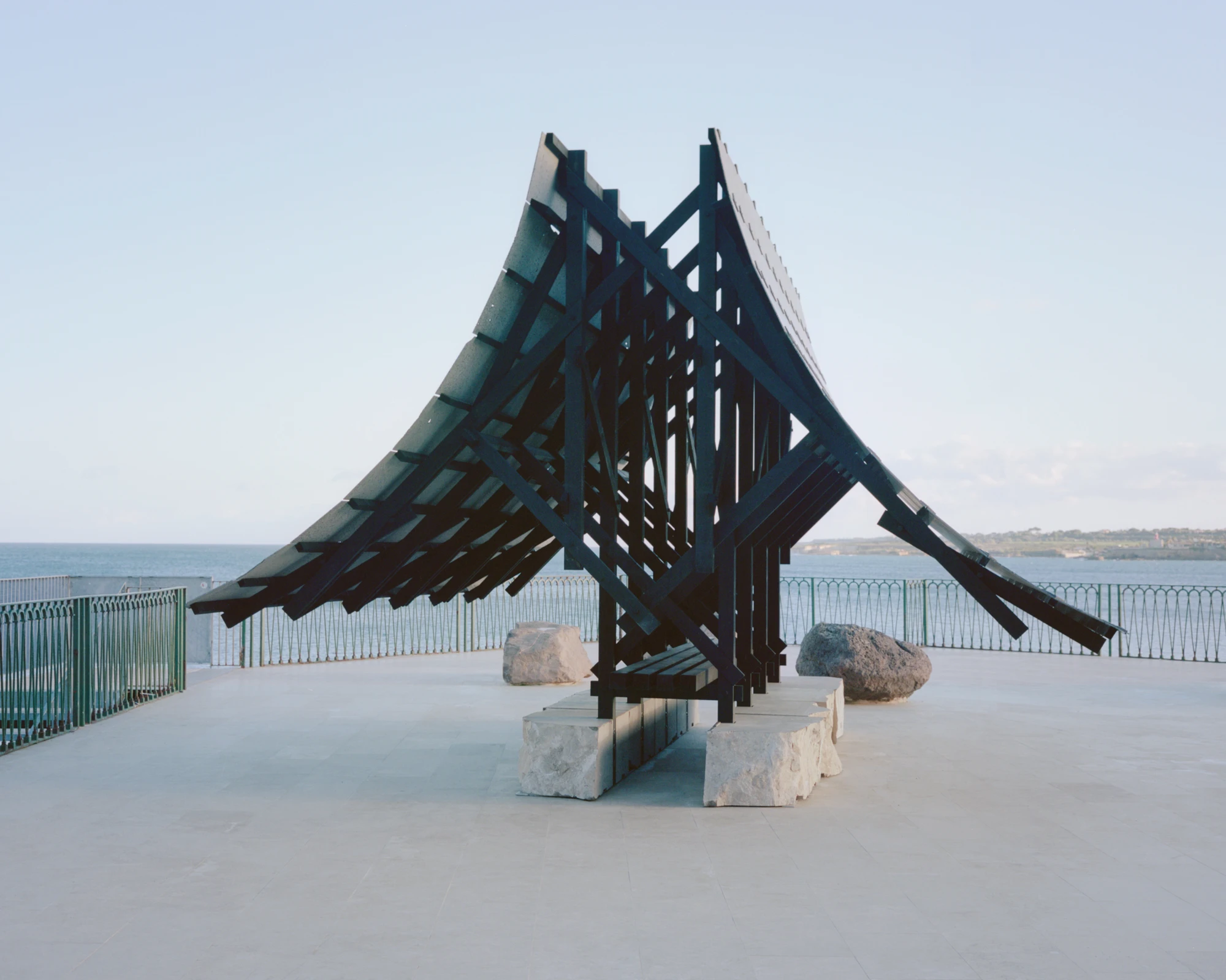 Pantalica Speculations by Leopold Banchini, An asymptotic timber and stone pavilion on a seaside promenade overlooking the Mediterranean, dark wood frame supporting stone-tile roof on massive limestone blocks