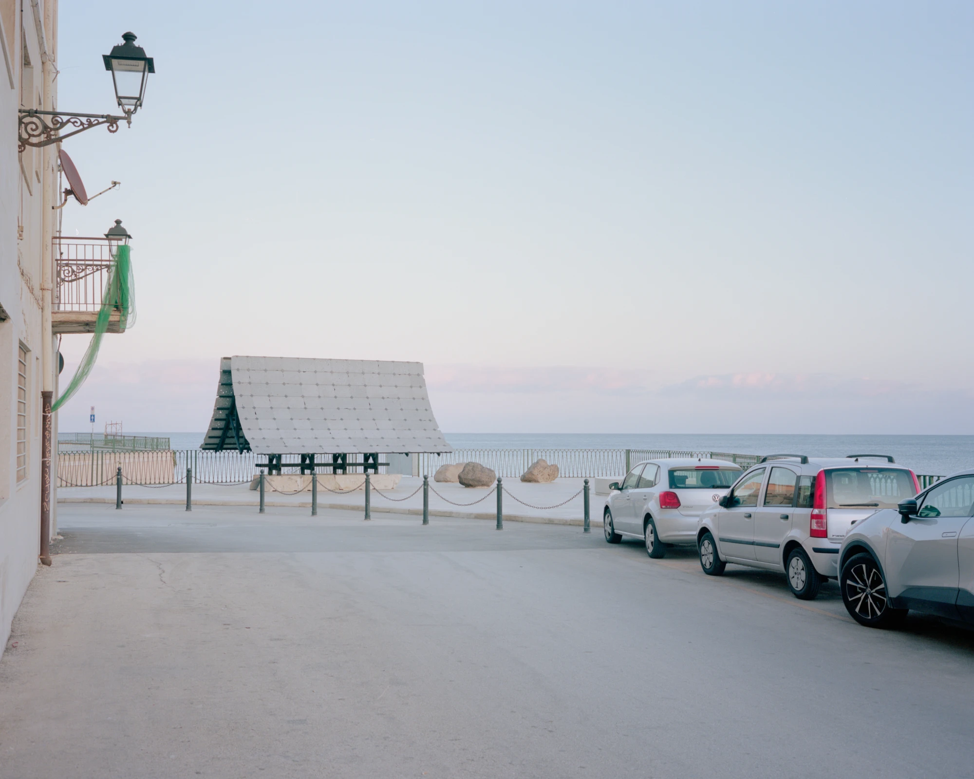 Pantalica Speculations by Leopold Banchini, An asymptotic timber and stone pavilion on a seaside promenade overlooking the Mediterranean, dark wood frame supporting stone-tile roof on massive limestone blocks