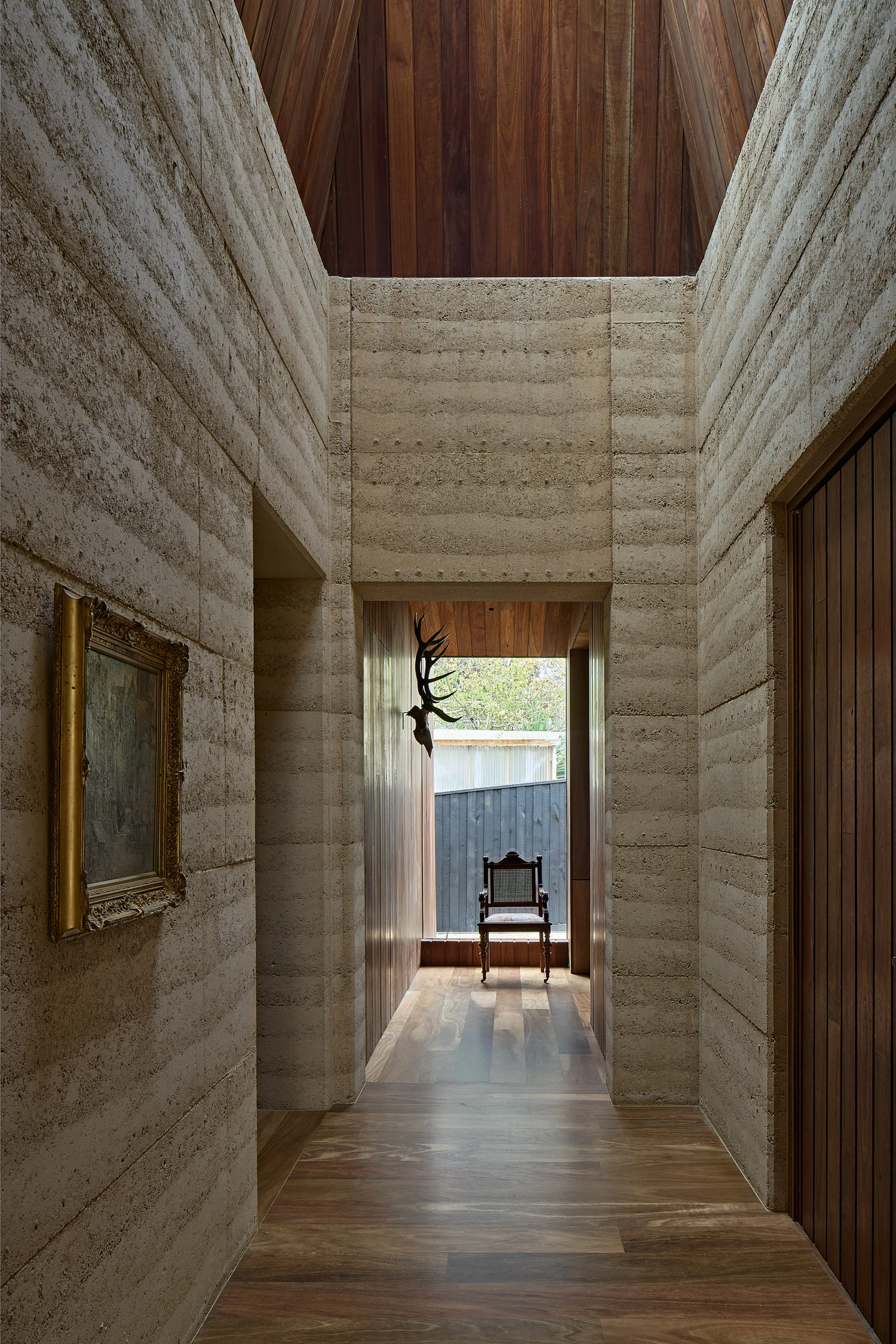 Homestead House by MRTN Architects — A corridor framed by rammed-earth walls with visible horizontal compaction striations extends toward a timber-lined threshold.