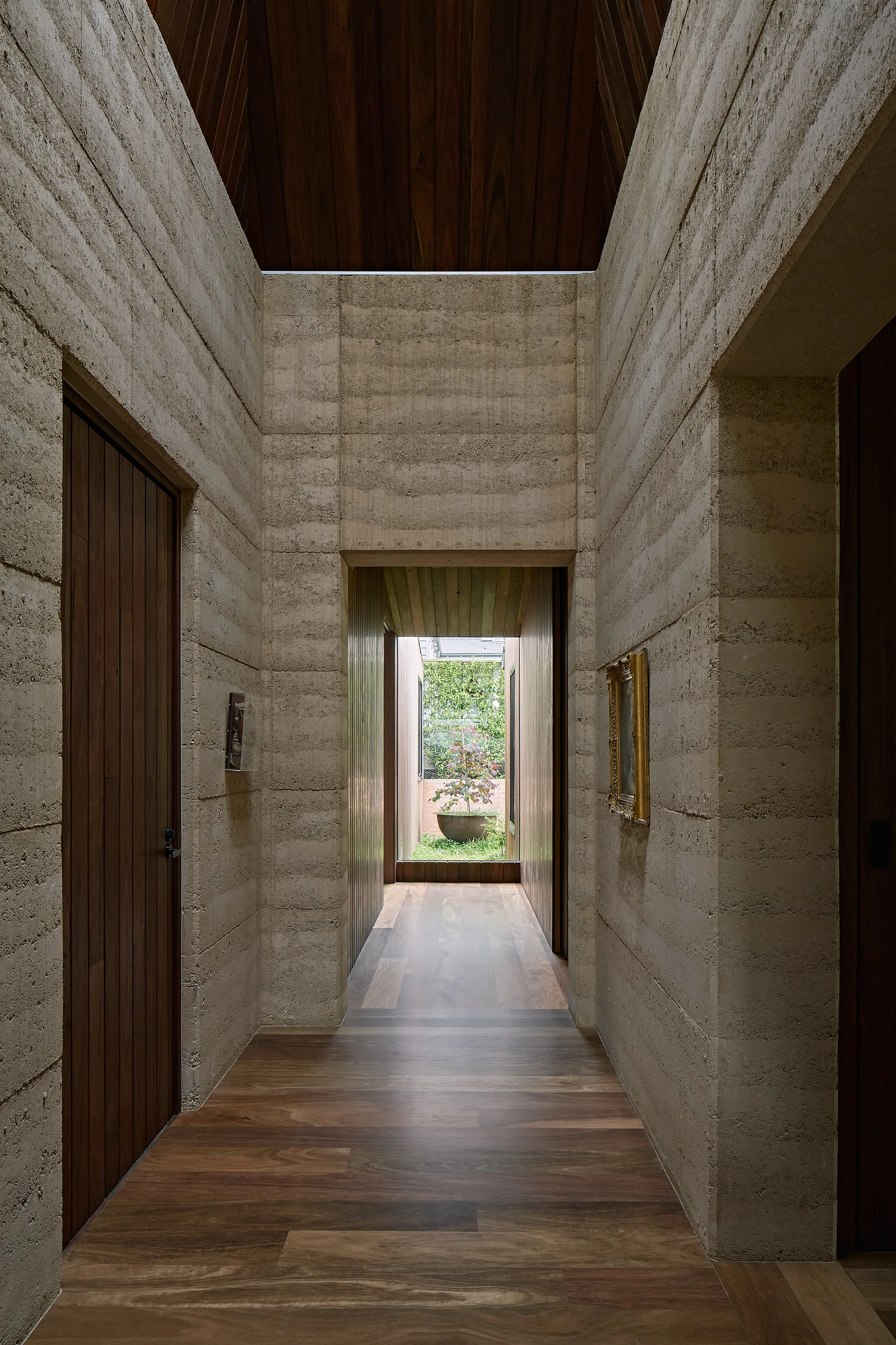 Homestead House by MRTN Architects — A narrow corridor framed by rammed-earth walls with visible horizontal compaction striations extends toward a threshold of diffused natural light.