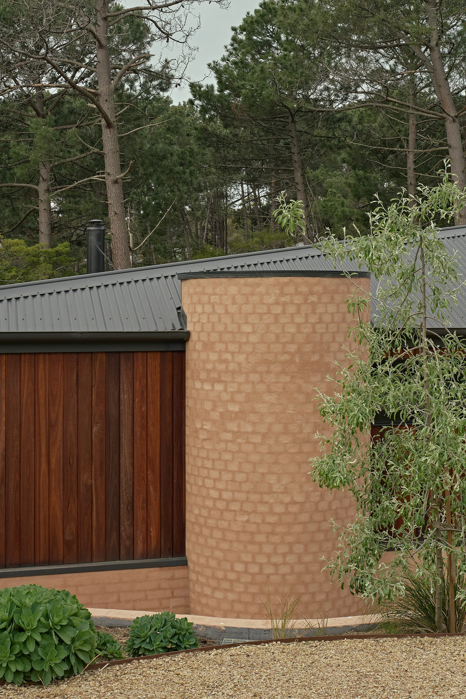 Homestead House by MRTN Architects — A curved rammed-earth wall dominates the foreground, its horizontal compaction striations visible across the terracotta surface.
