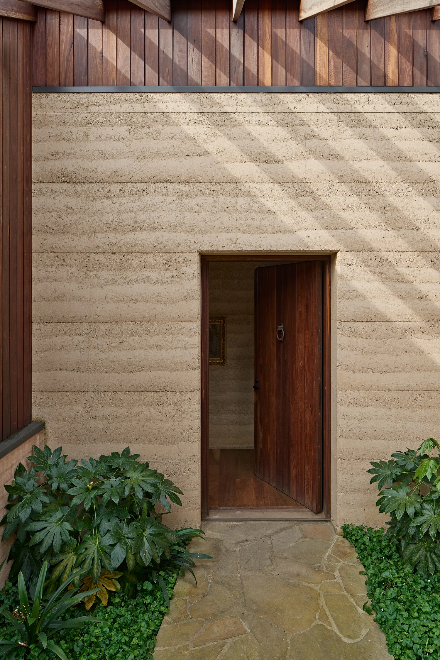 Homestead House by MRTN Architects — A rammed-earth wall rendered in pale ochre dominates the frame, its horizontal compaction striations catching afternoon light in sharp relief.