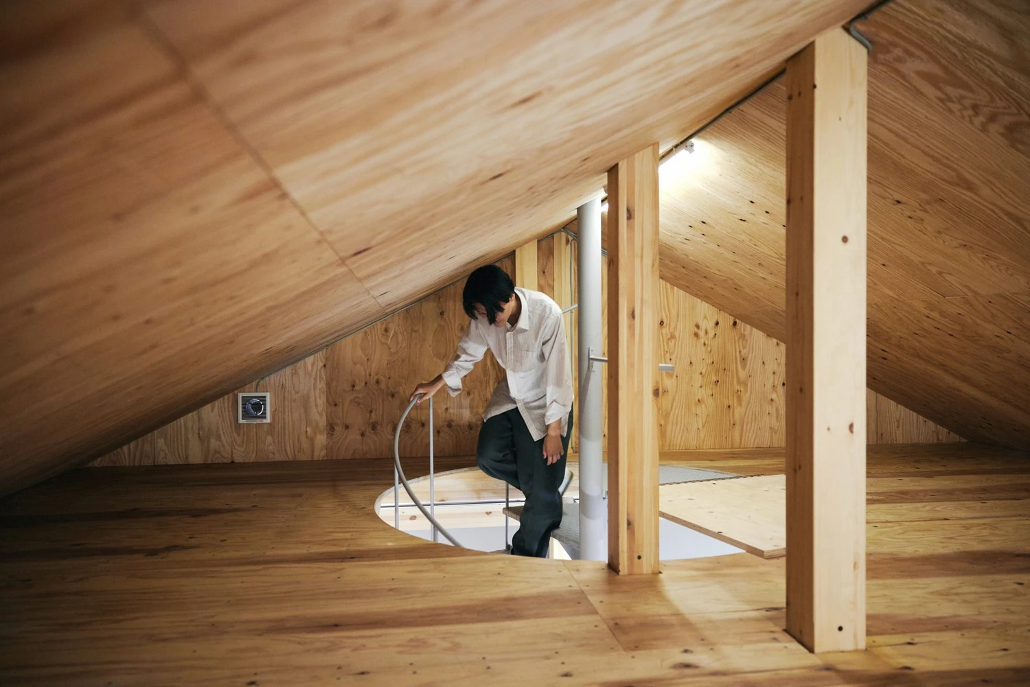 Uchida Shoten Head Office by Jo Nagasaka / Schemata Architects — Interior view of the Uchida Shoten head office attic space, showing a man in white shirt and dark trousers descending through a circular aperture in the...