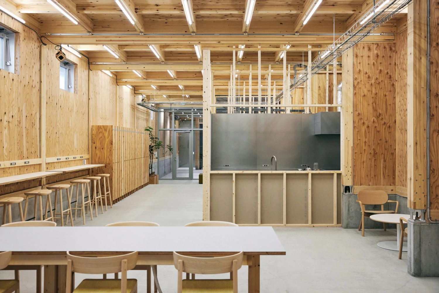 Uchida Shoten Head Office by Jo Nagasaka / Schemata Architects — Interior view of a timber-clad open-plan space reveals the post-and-beam structural logic beneath pitched timber ceilings lined with fluorescent strips.