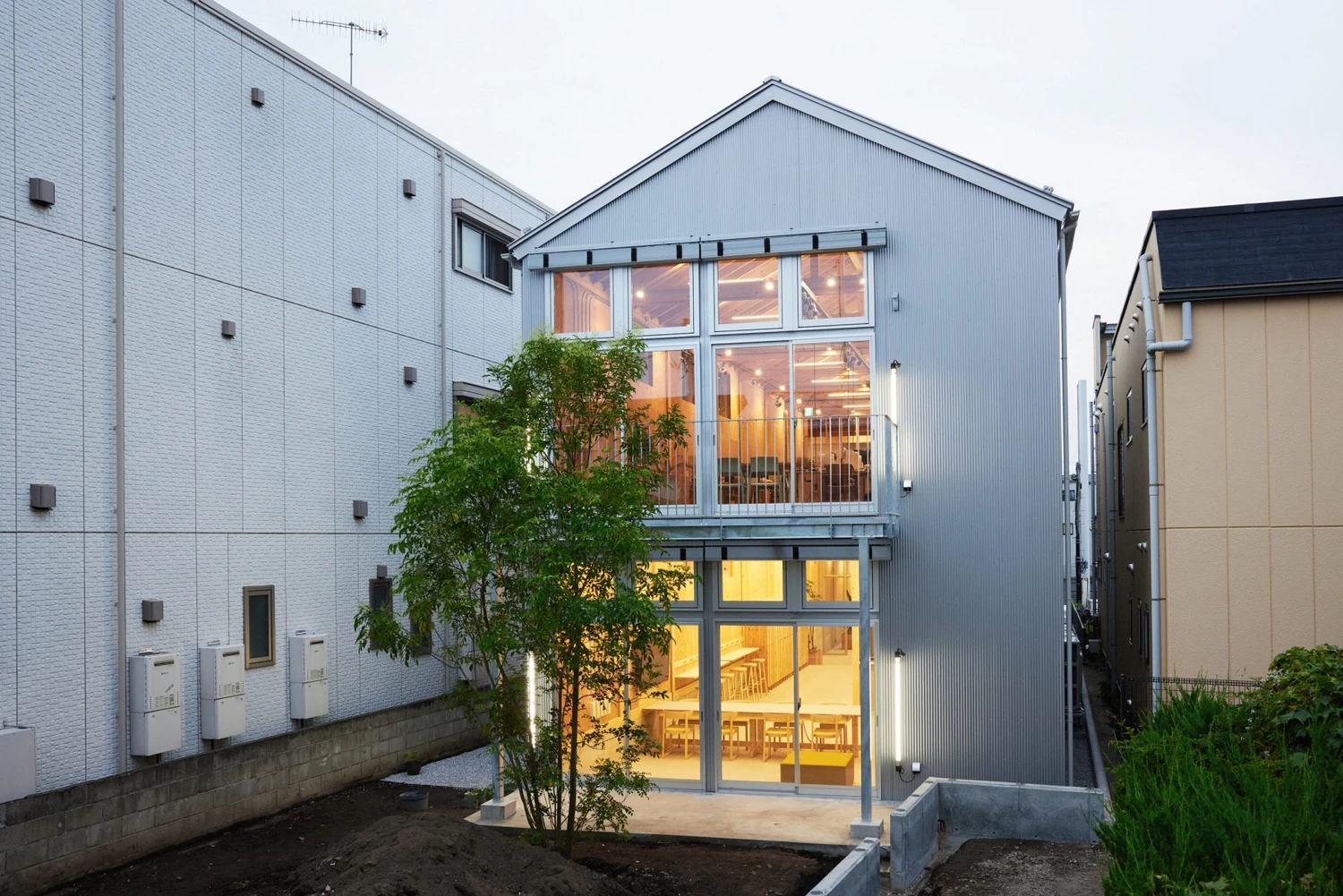 Uchida Shoten Head Office by Jo Nagasaka / Schemata Architects — Evening light spills through a two-story gridded window assembly, illuminating the timber interior of this compact Fujisawa office building.