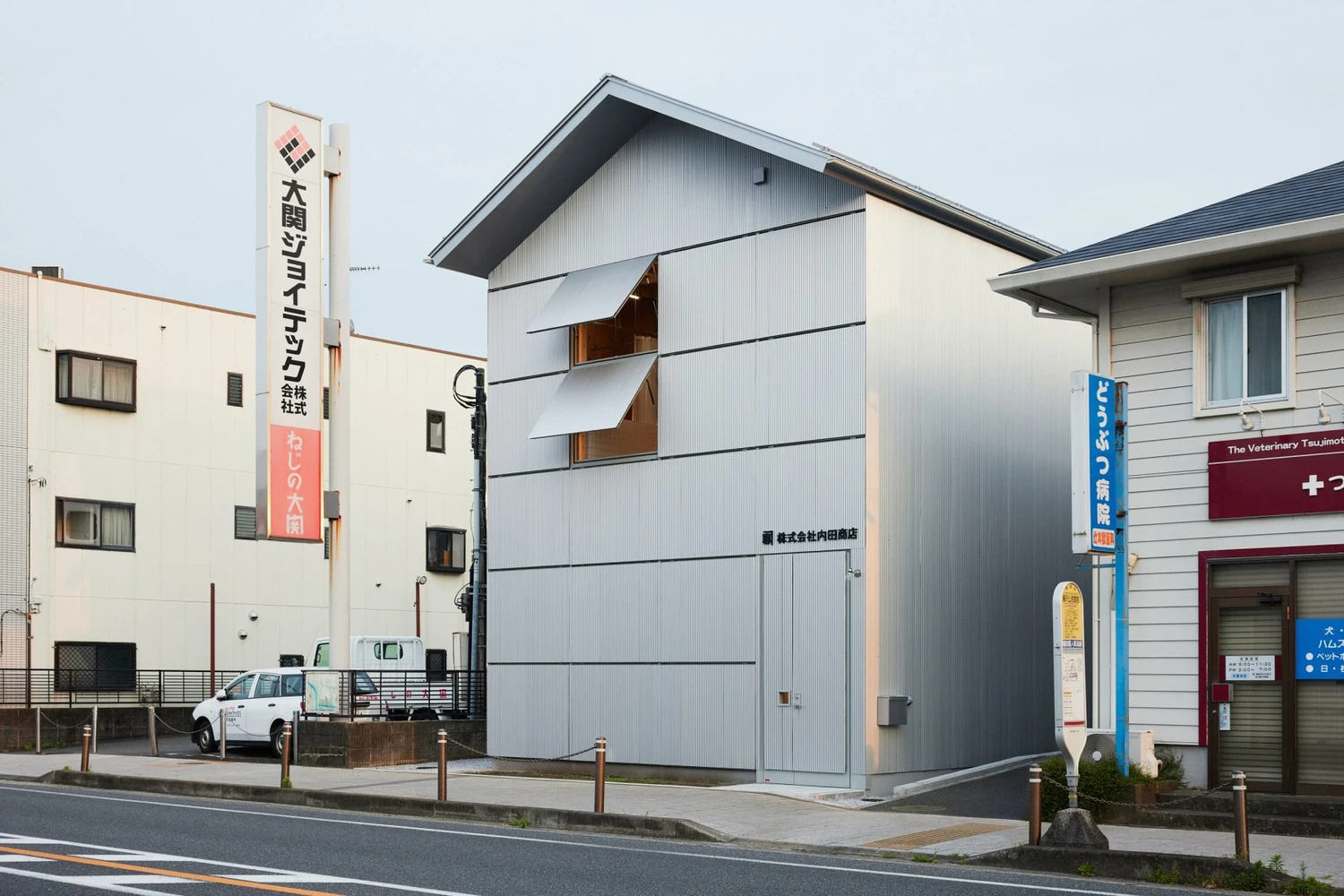 Uchida Shoten Head Office by Jo Nagasaka / Schemata Architects — A three-story corrugated metal structure with white cladding occupies a narrow corner plot in Fujisawa, its form echoing the pitched-roof vernacular of...