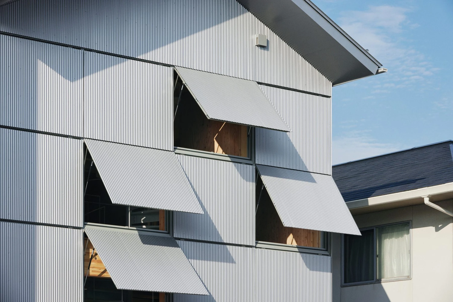 Uchida Shoten Head Office by Jo Nagasaka / Schemata Architects — Diagonal hinged metal awning windows dominate the corrugated white metal facade, each panel angled to reveal warm timber interiors and glass apertures beneath.