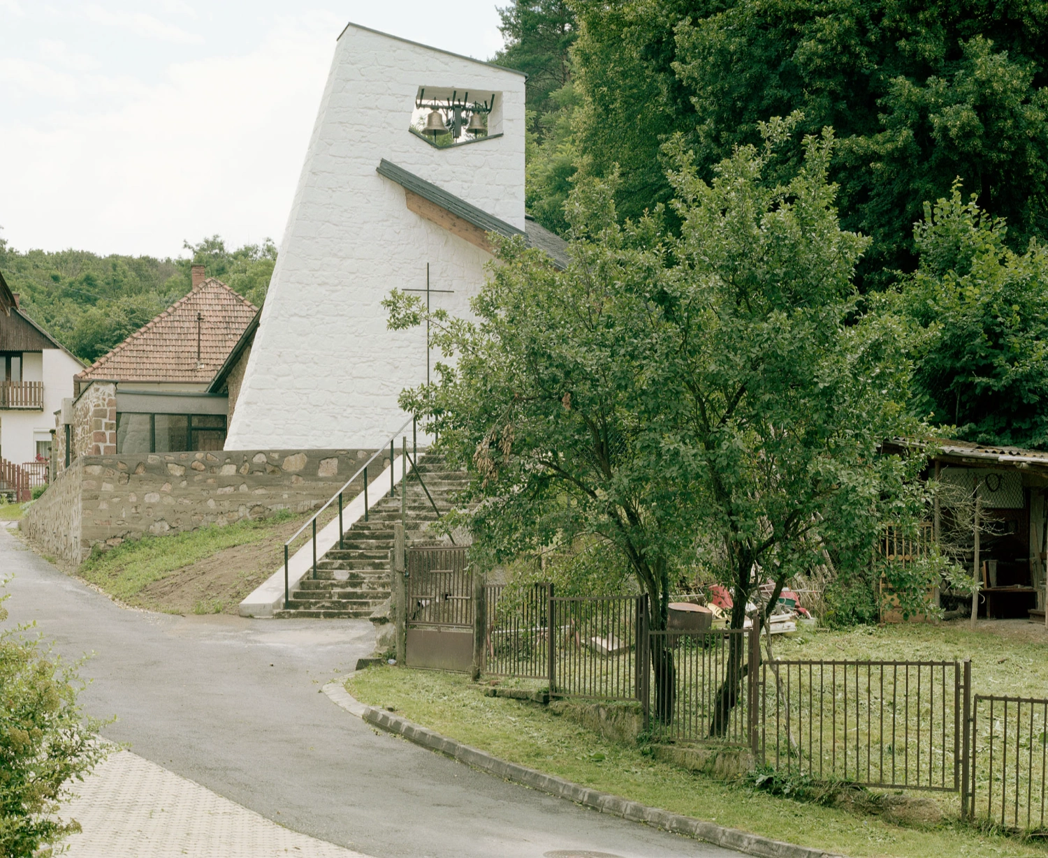 Restoration of the Church of Cserépváralja by Partizan Architecture — The restored church tower rises as a monolithic white rendered volume against dense forest backdrop, its inverted roofline creating a pronounced shadow line...