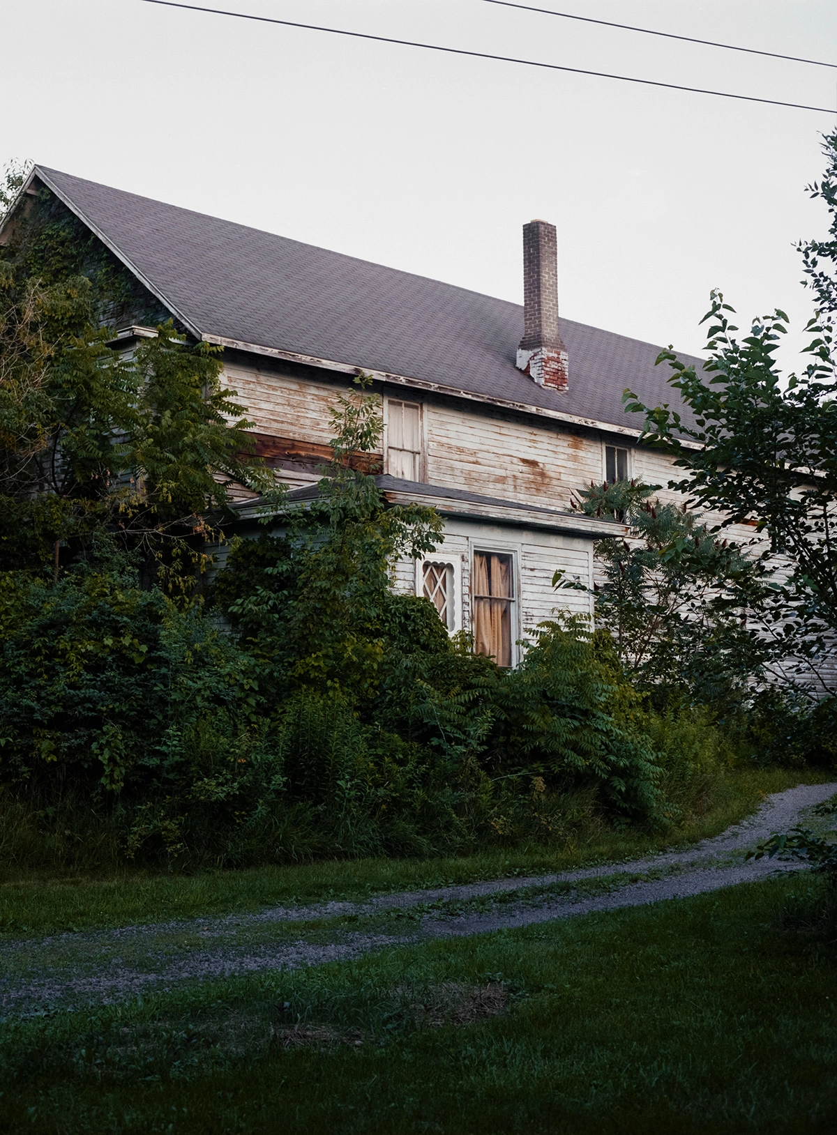 We Keep Swimming, Until We All Reach Home by Jillian Guyette — A two-story residential structure with weathered wood siding and a dark pitched roof punctuated by a brick chimney.