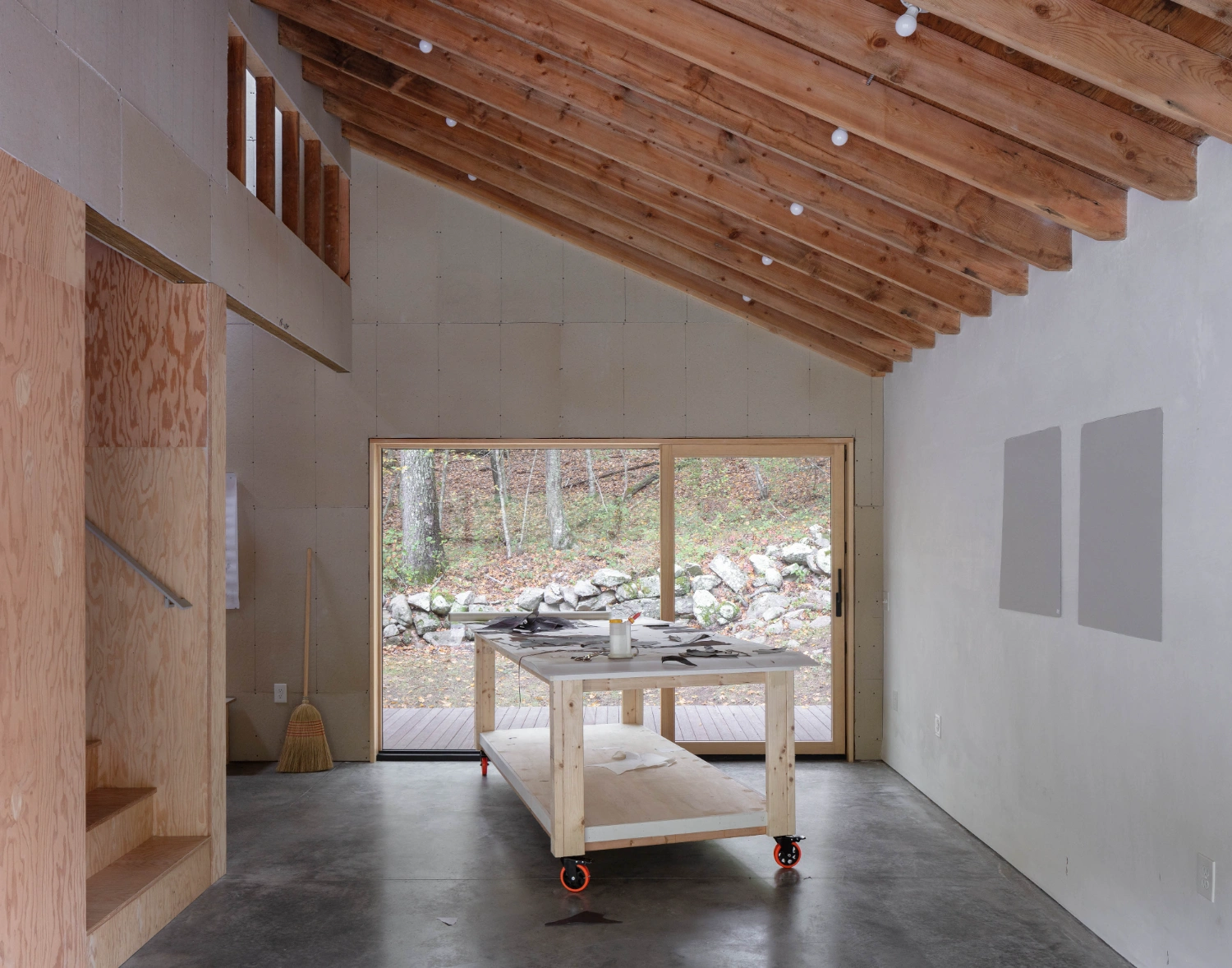 Studio Barn by Worrell Yeung — A wooden workbench on red casters occupies the center of the studio's open floor plan, positioned between the entrance and large glazed doors that frame the...