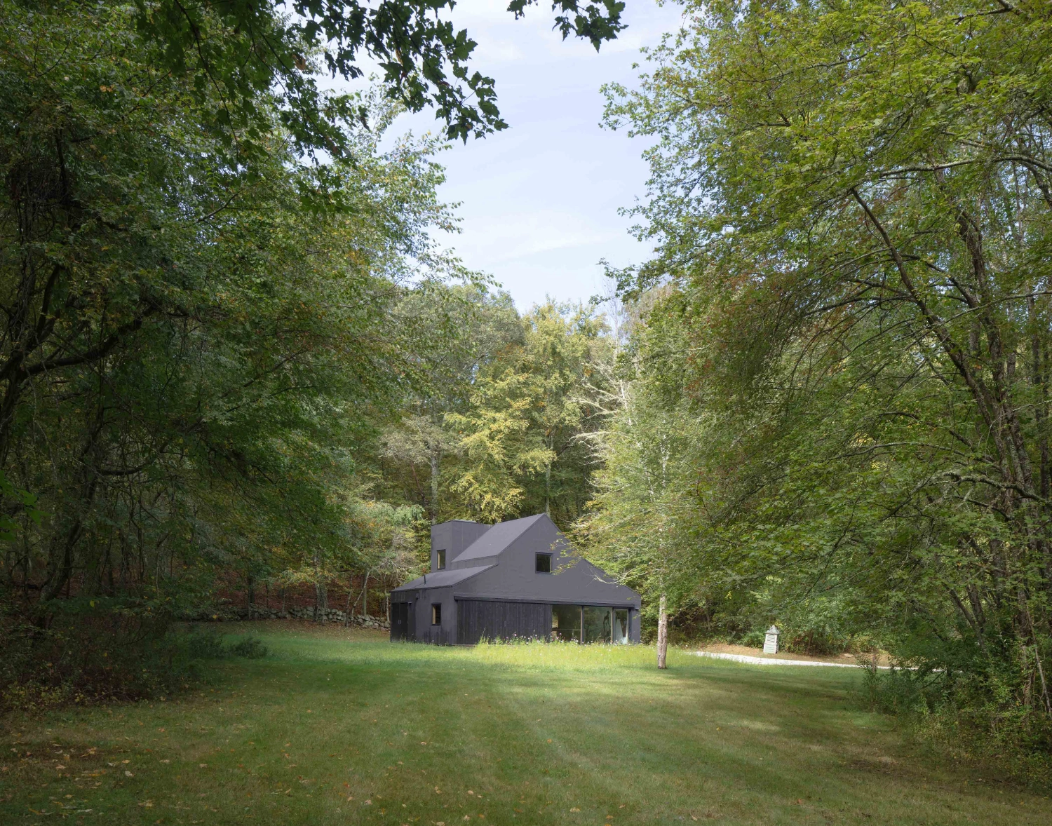 Studio Barn by Worrell Yeung — A dark-clad residential structure sits within a clearing surrounded by dense deciduous forest.