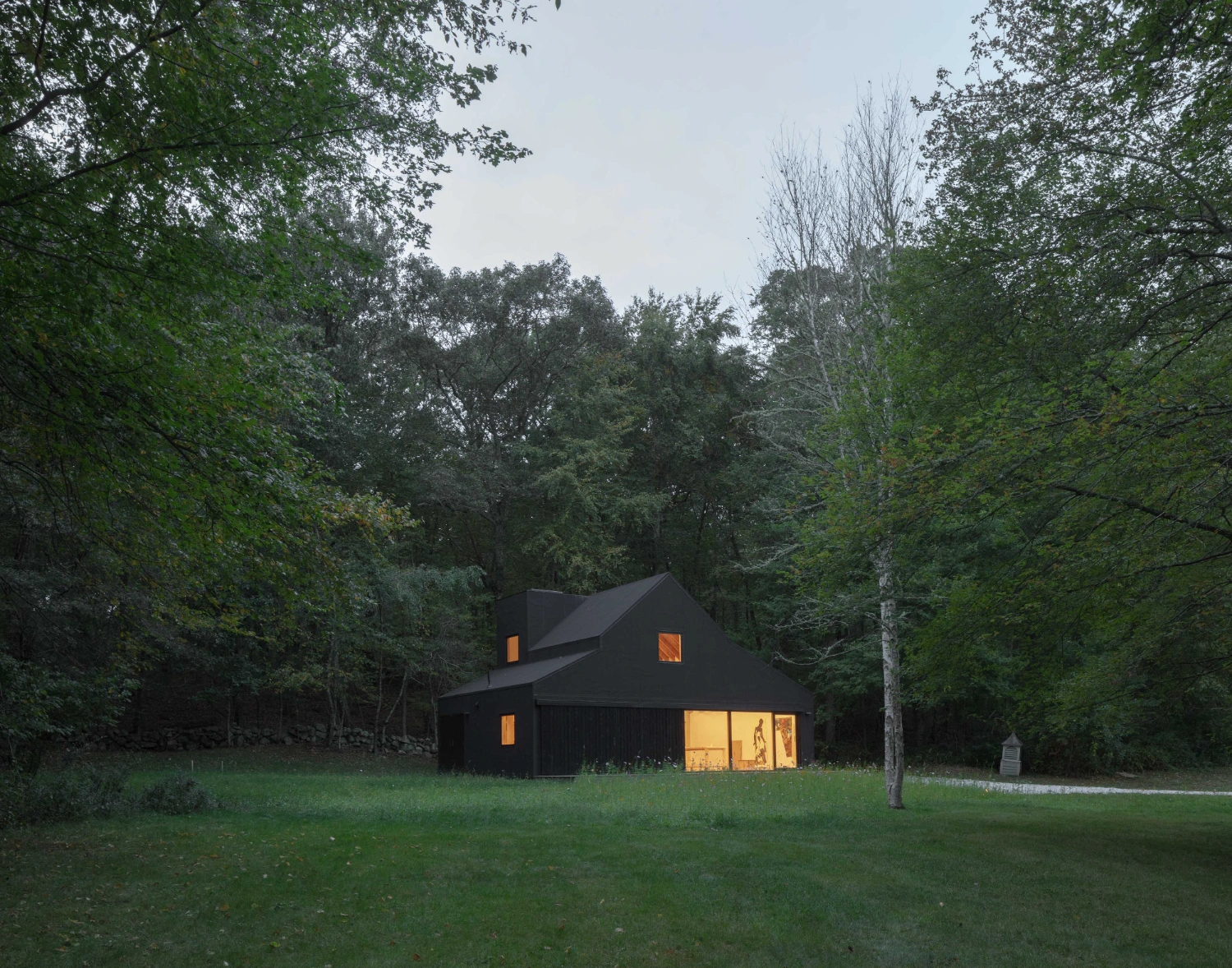 Studio Barn by Worrell Yeung — A black-clad barn structure emerges from a clearing in dense woodland, its geometric form defined by a steeply pitched roof and clean rectangular volumes.