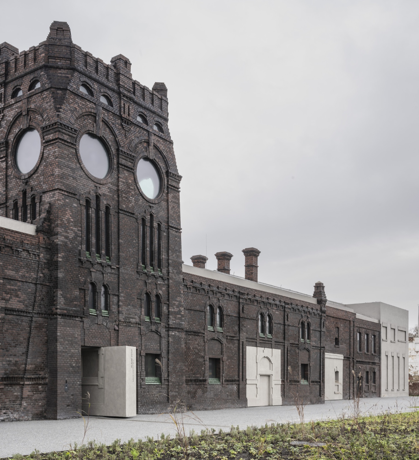 PLATO Contemporary Art Gallery by KWK PROMES — The pavilion's geometric concrete volume punctures the brick facade, its sharp vertical edges creating a severe material contrast against the weathered masonry.
