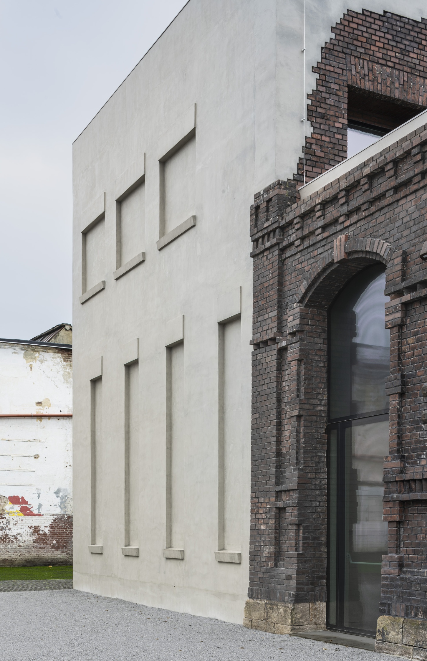 PLATO Contemporary Art Gallery by KWK PROMES — A monolithic concrete pavilion with recessed vertical window openings inserted into a dark brick industrial structure.