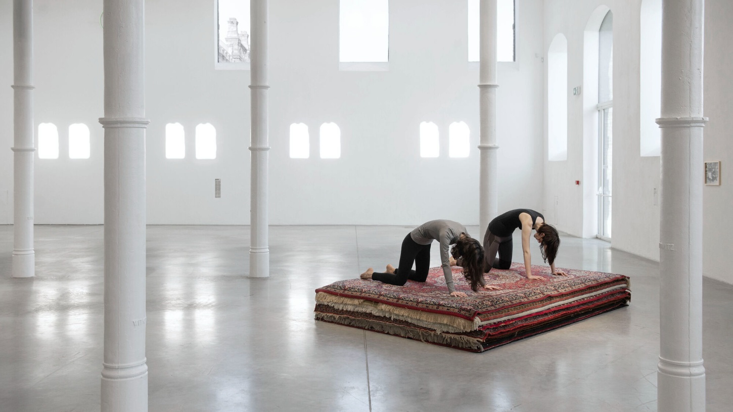 PLATO Contemporary Art Gallery by KWK PROMES — Two figures perform a synchronized yoga pose on layered traditional rugs within the gallery's minimalist interior.