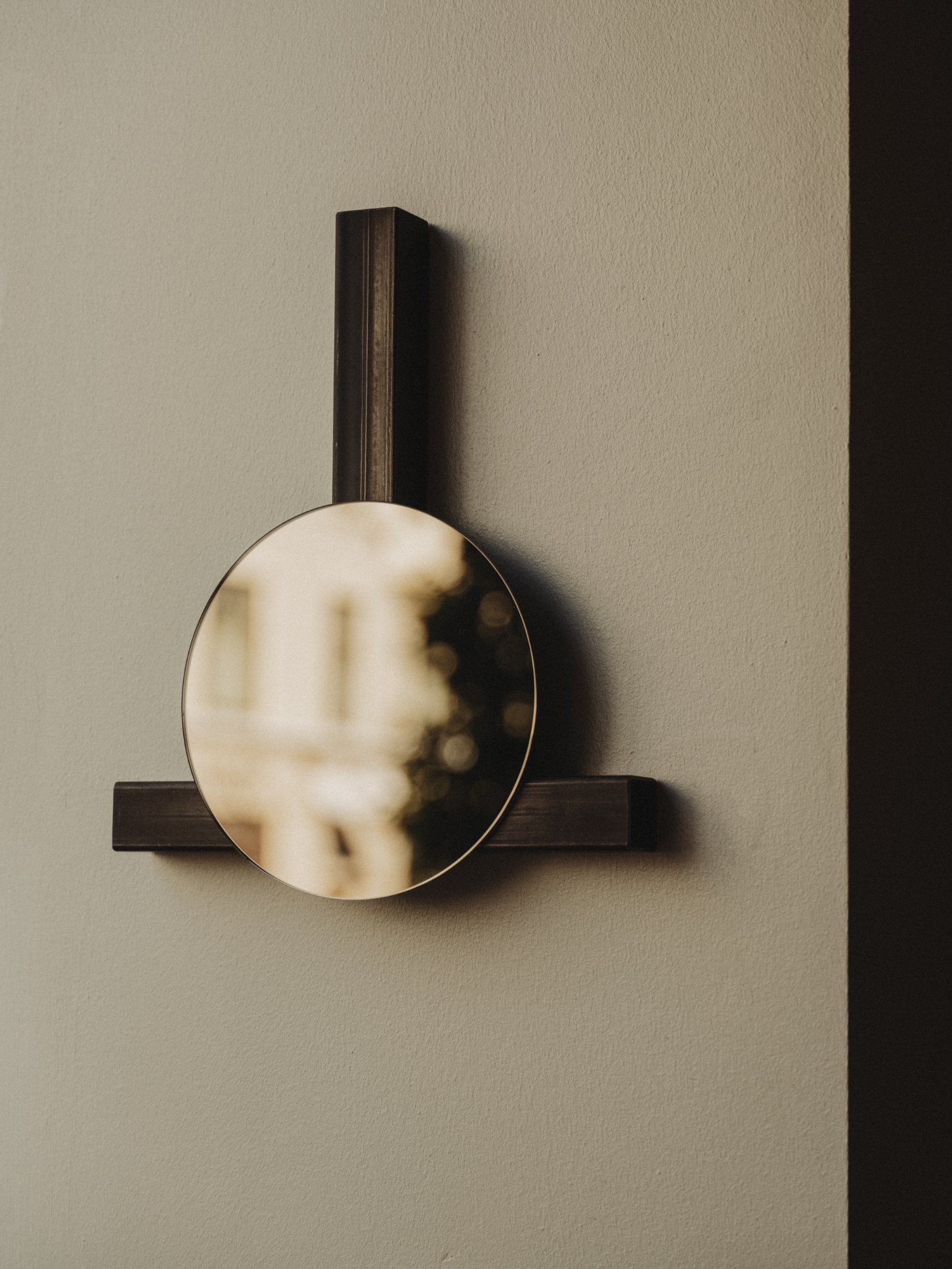 Amator Restaurant by wiercinski-studio — A circular mirror with a brass or bronze frame is mounted on a dark timber bracket that extends horizontally from a cream-colored wall.